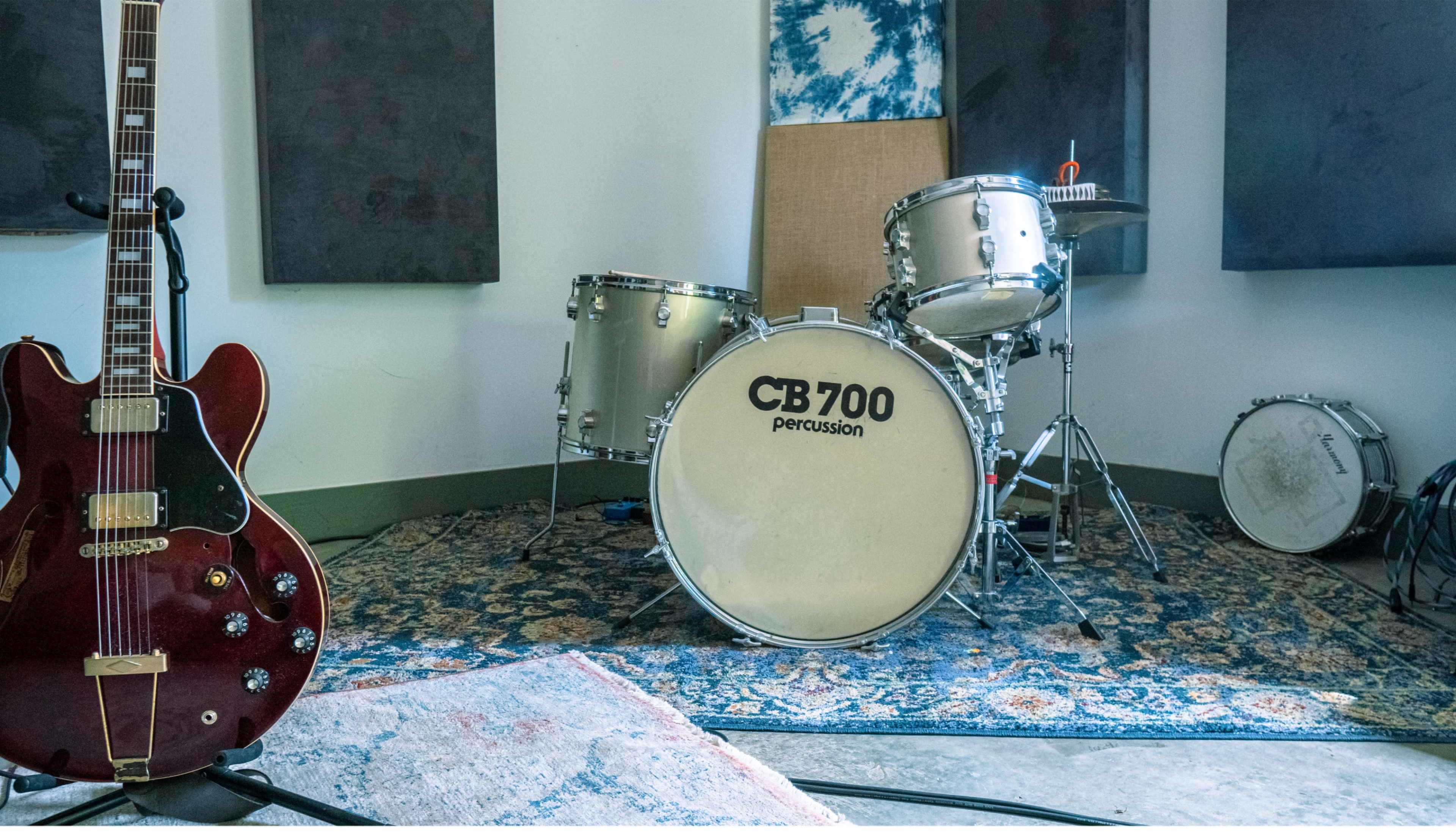 A red electric guitar stands next to a drum set in a music rehearsal studio.