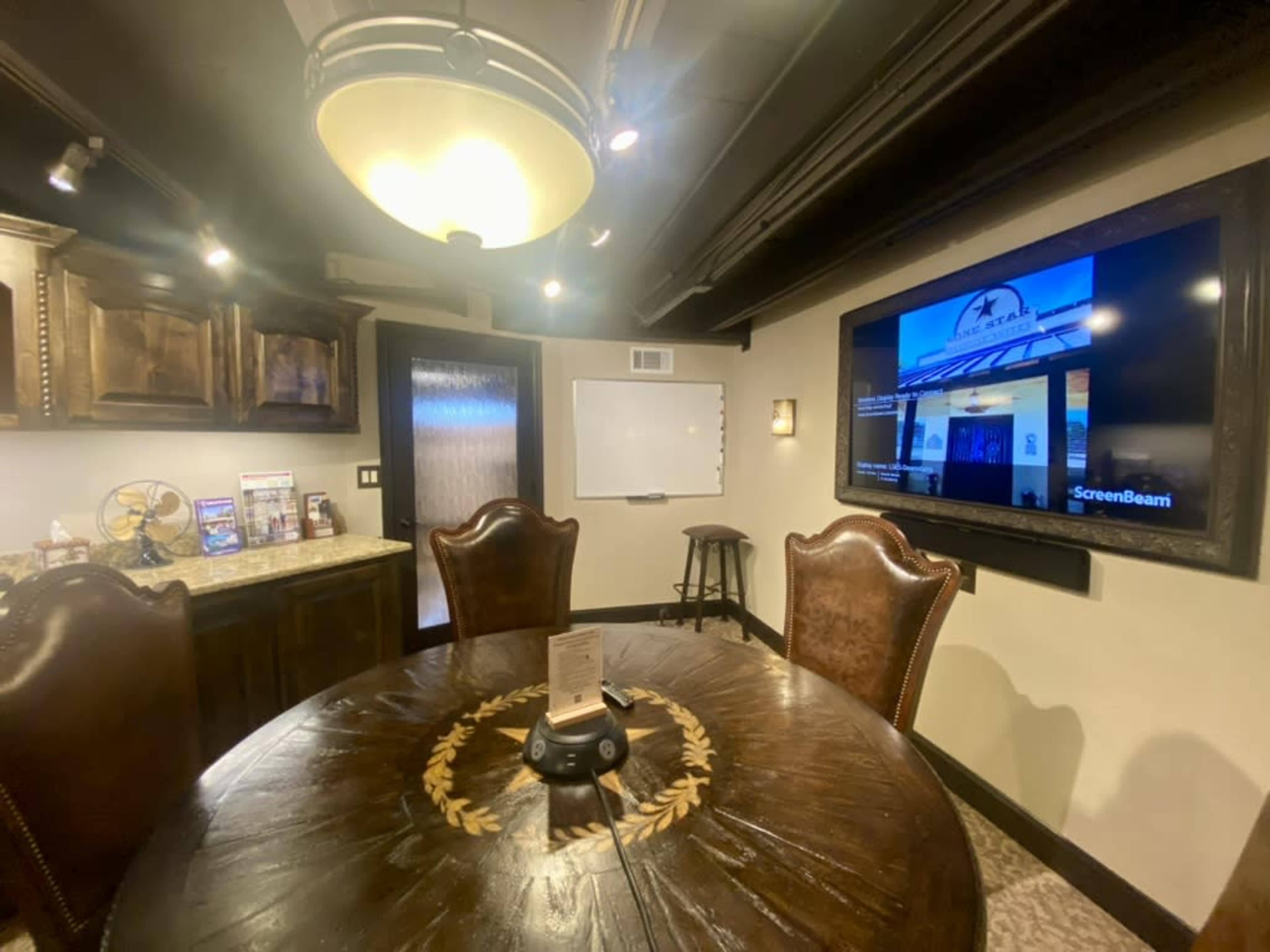 The image shows a conference room featuring a round wooden table, leather chairs, a flat-screen TV mounted on the wall, and a whiteboard.