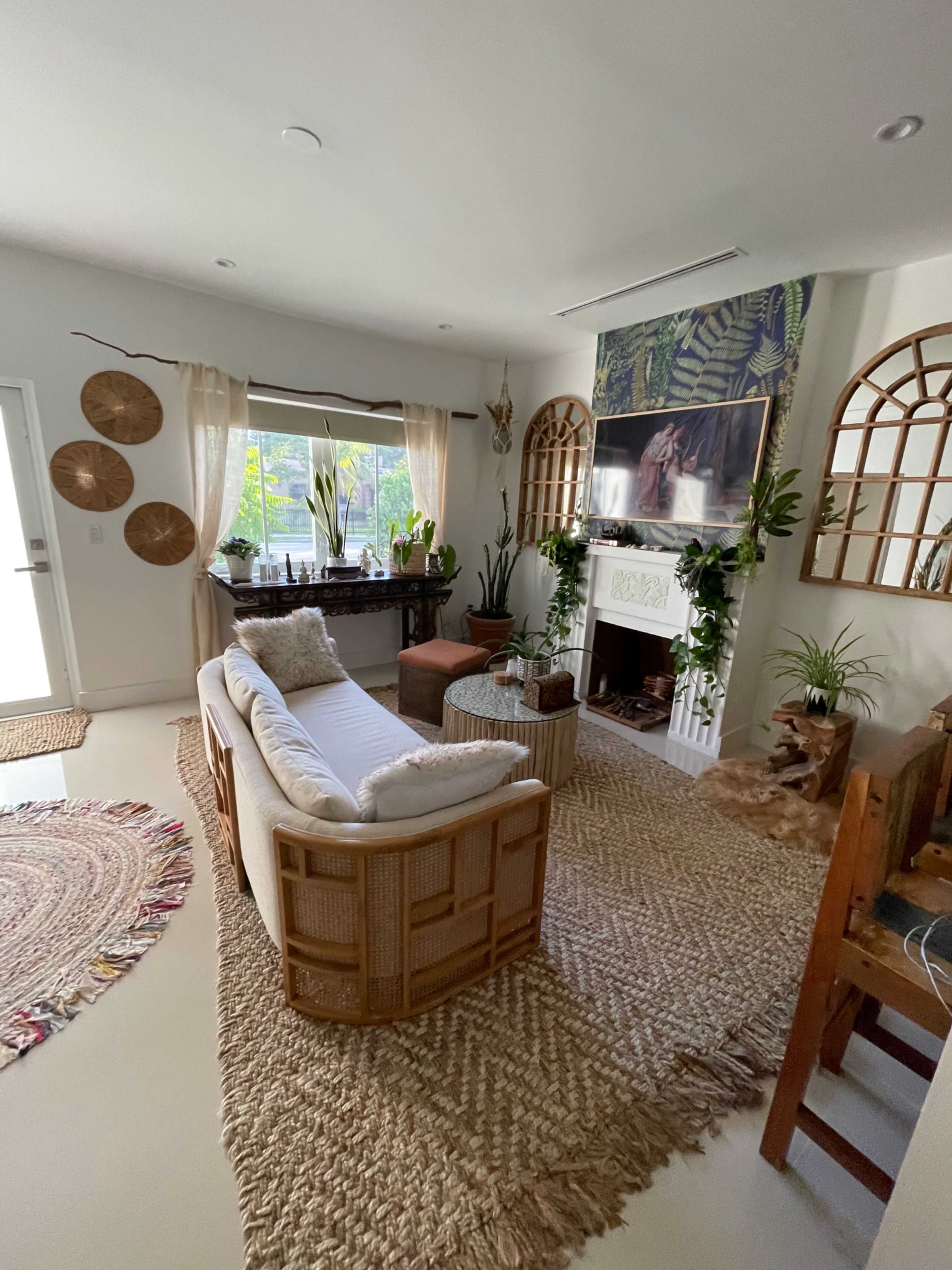 A cozy living room features a white sofa, a large TV mounted on the wall, and a mix of plants and decorative elements around a central coffee table.