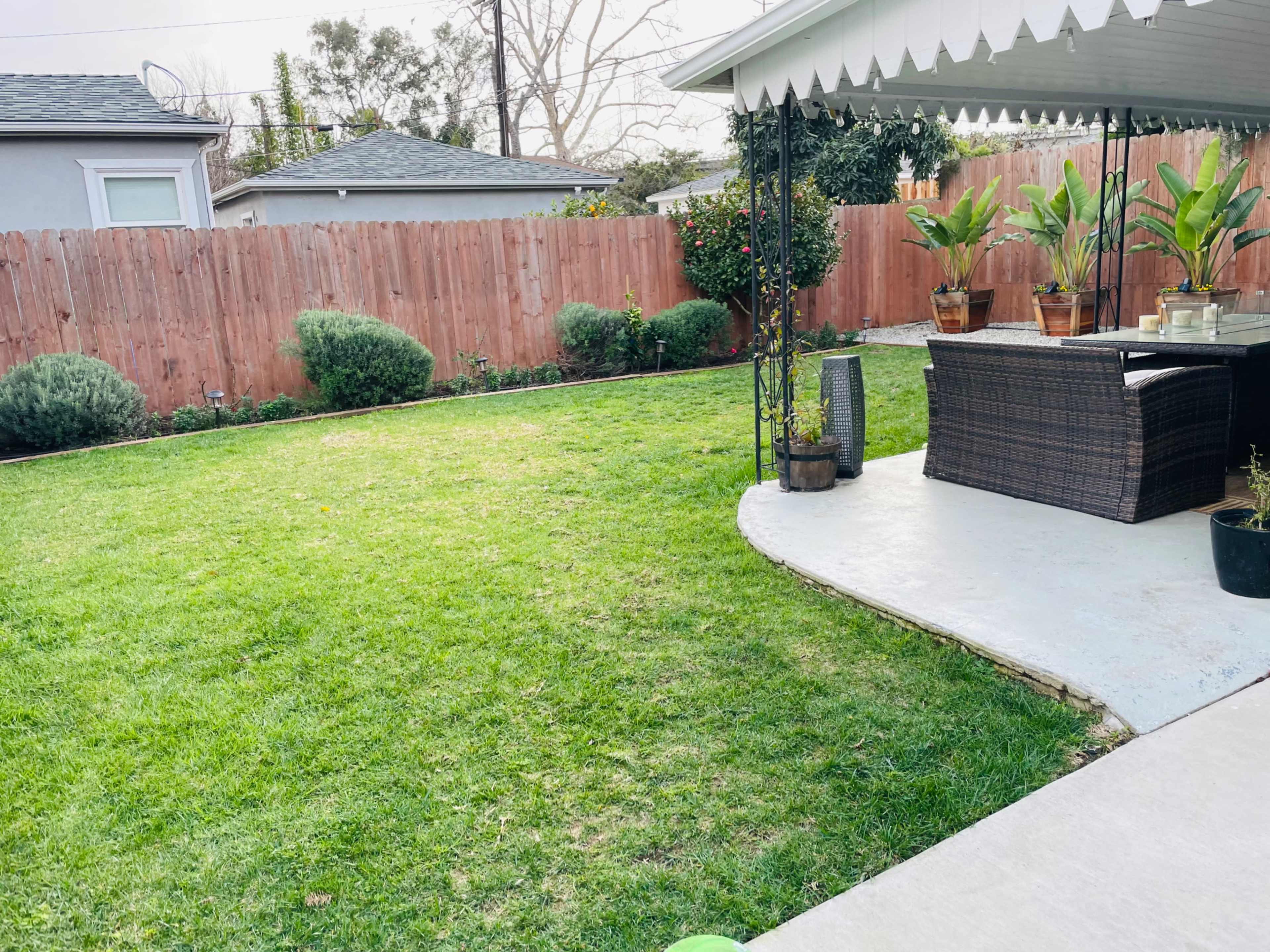 A grassy backyard with a patio area featuring potted plants and outdoor furniture, surrounded by a wooden fence.