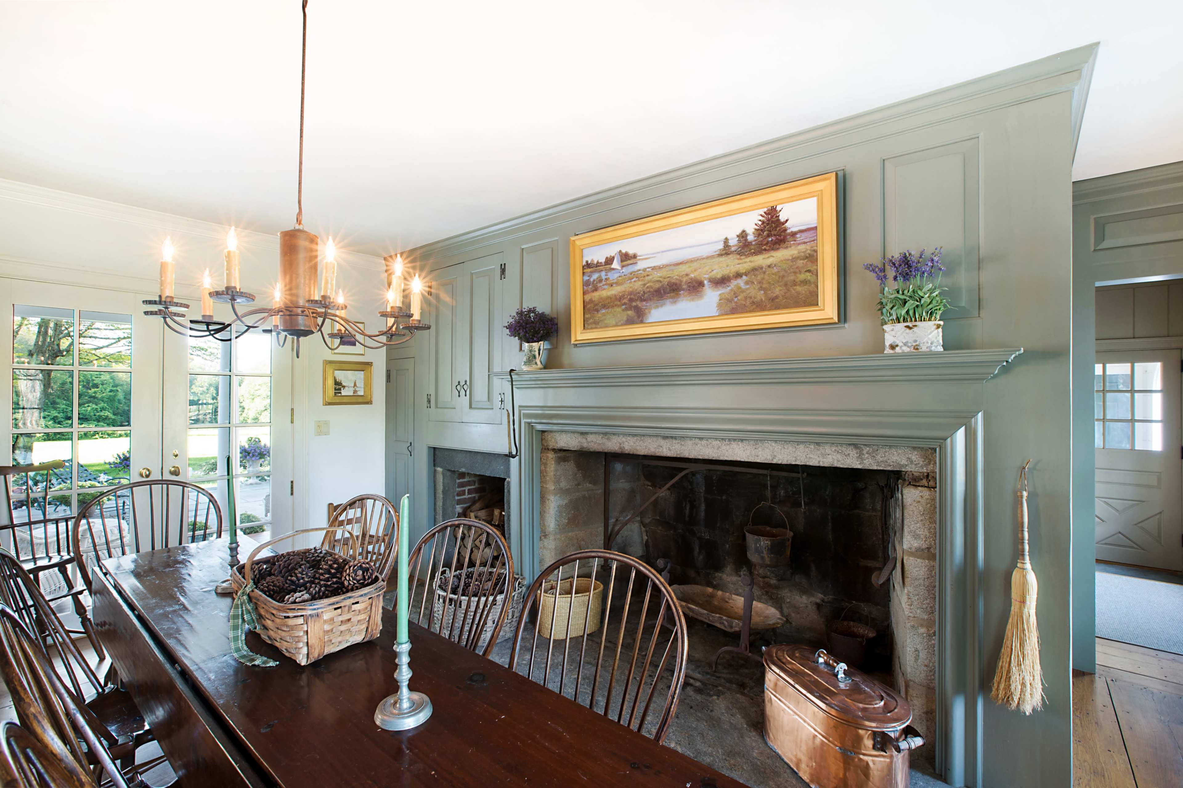 A dining room with a long wooden table, a chandelier, a stone fireplace, and a large painting above the mantel.
