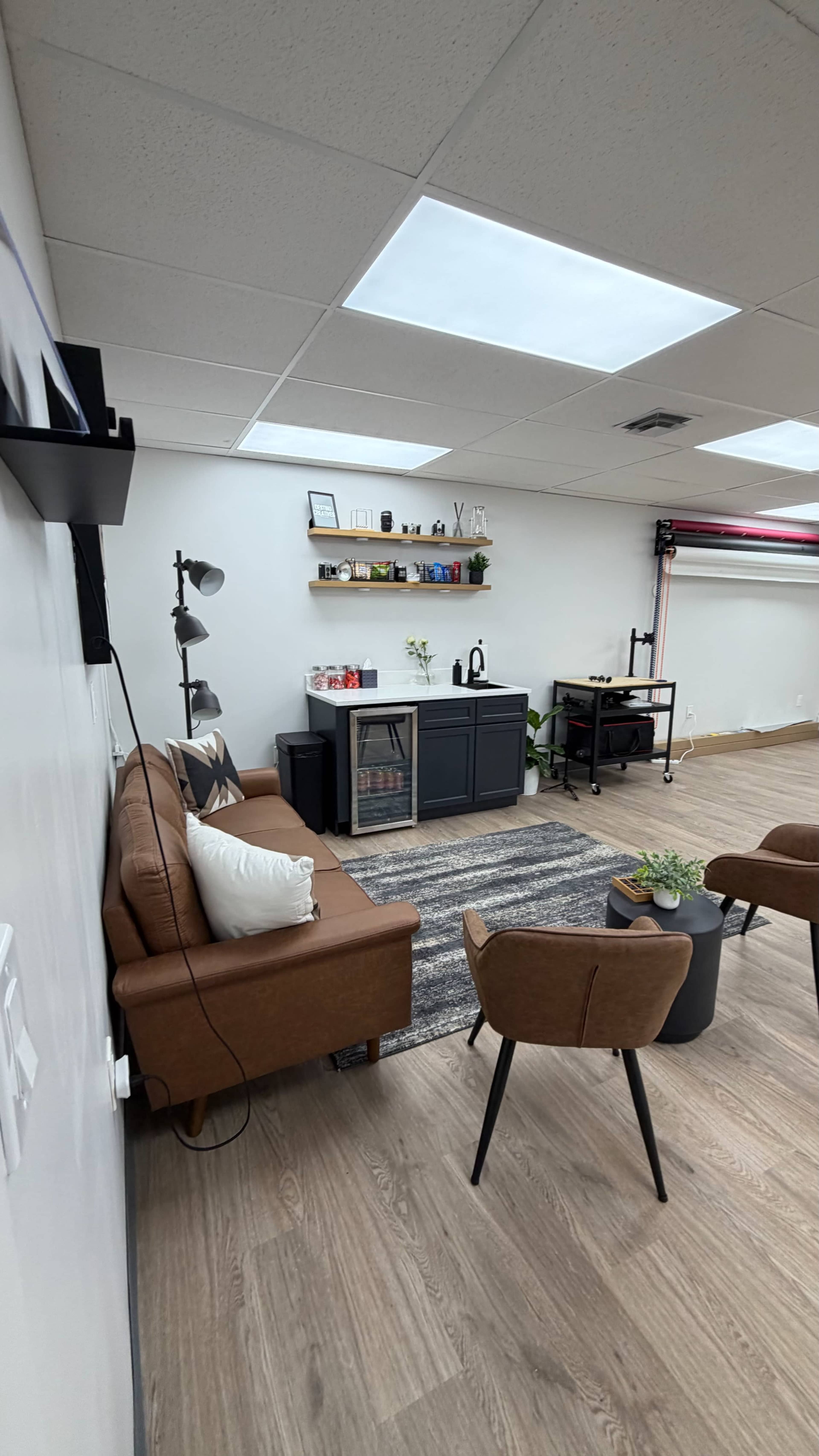 The image shows a modern office space featuring a brown sofa, two chairs, a coffee table, and a wall-mounted shelf with decor items.