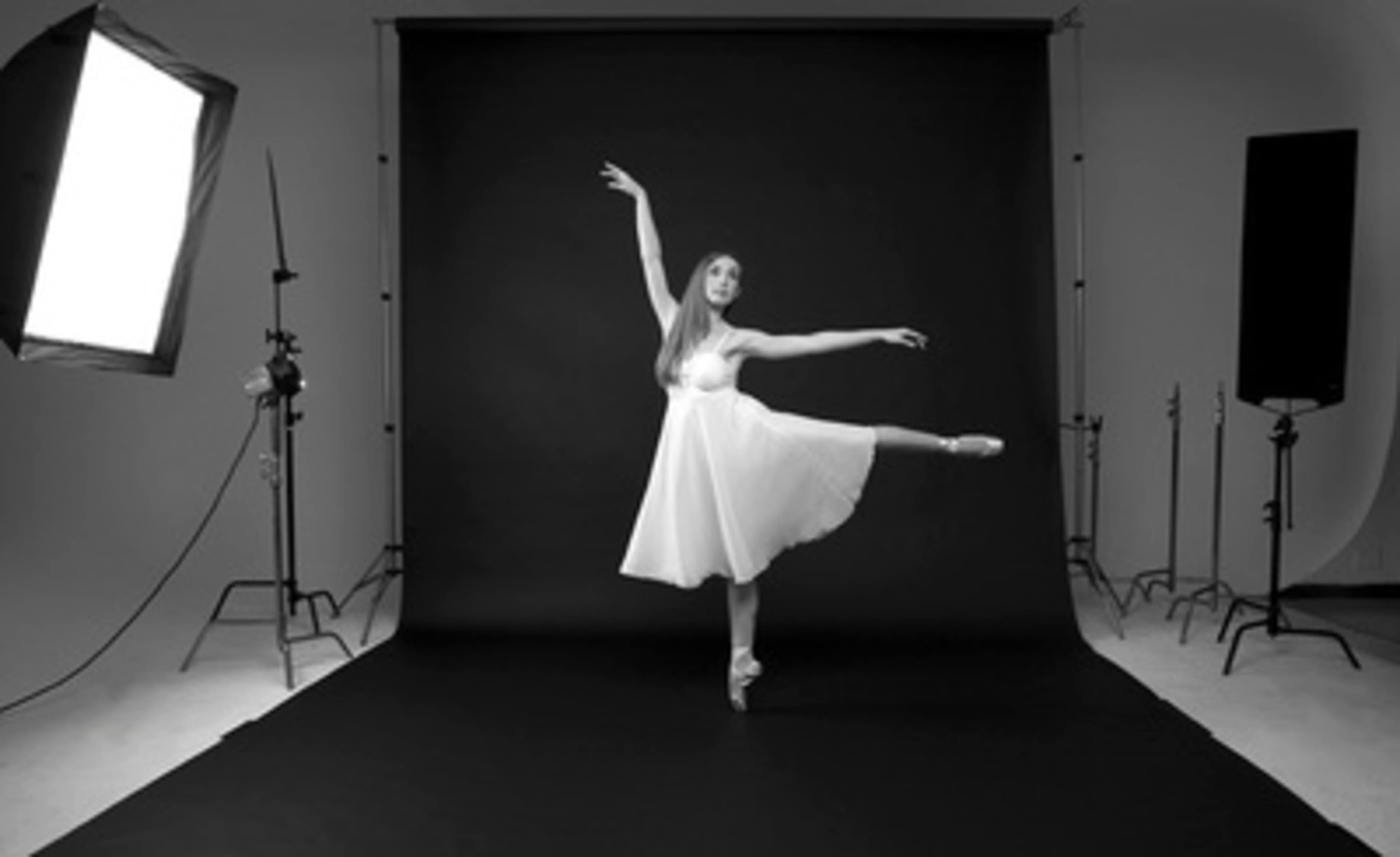 A dancer in a white dress performs an arabesque on a black stage surrounded by studio lighting equipment.