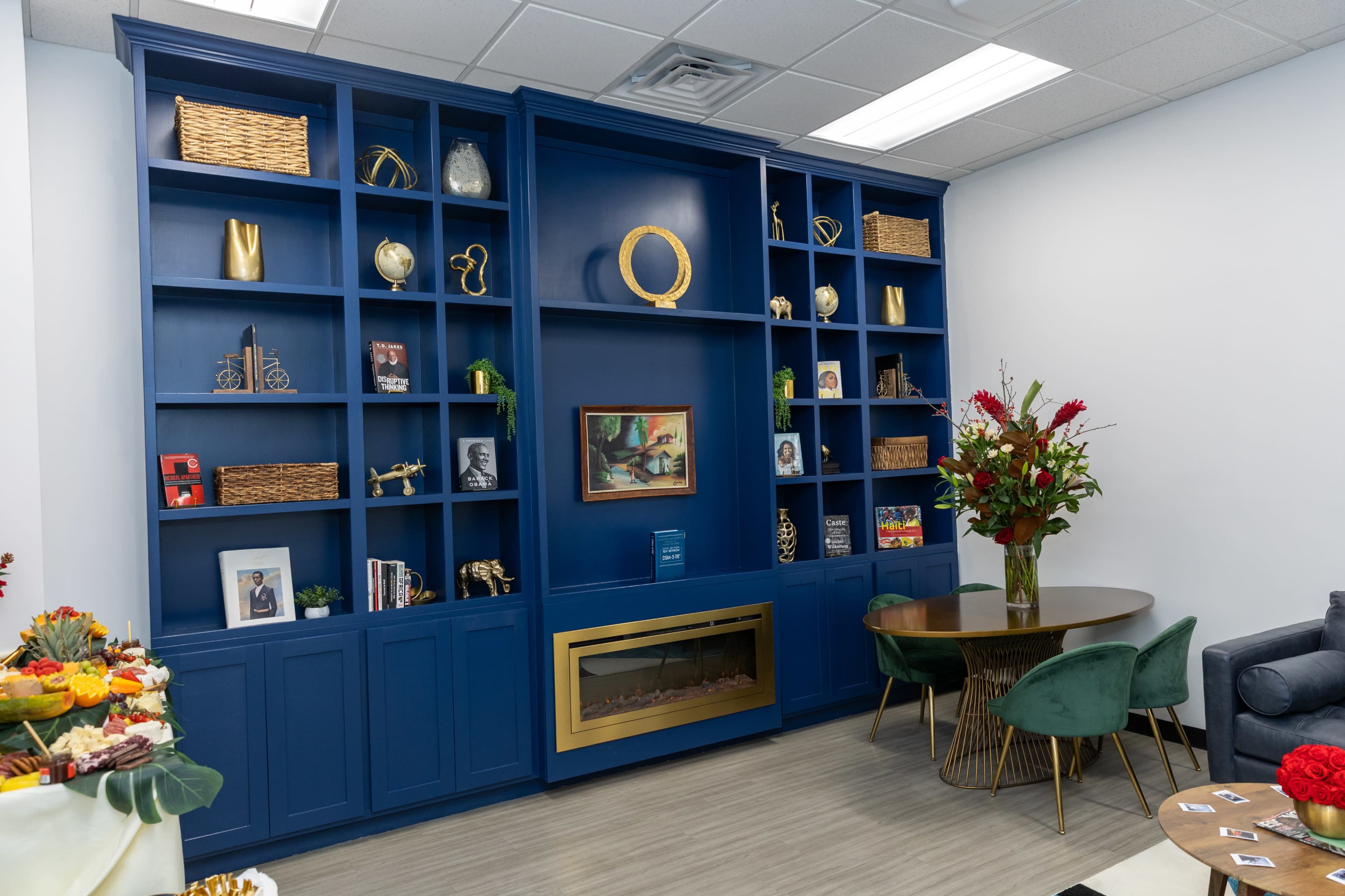 The image shows a modern room with a blue bookshelf filled with decorative items, a round dining table with green chairs, and a faux fireplace beneath the shelf.