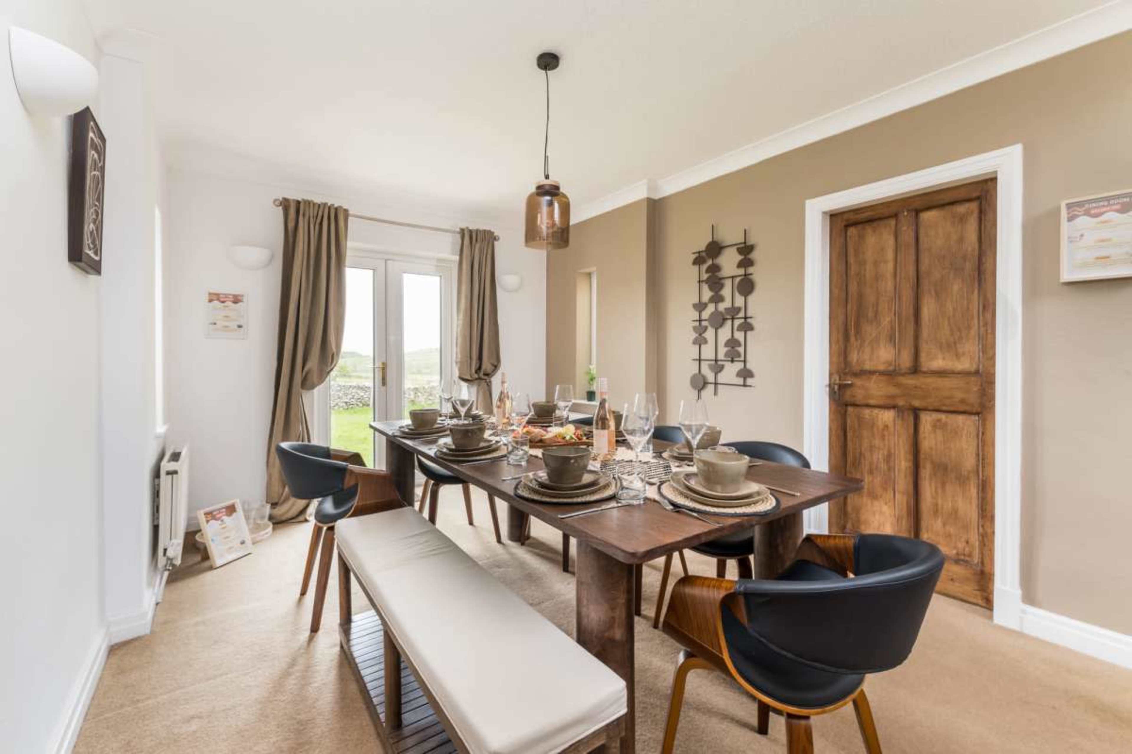 The dining room features a wooden table set with dinnerware and surrounded by various chairs, with a large window allowing natural light to enter.