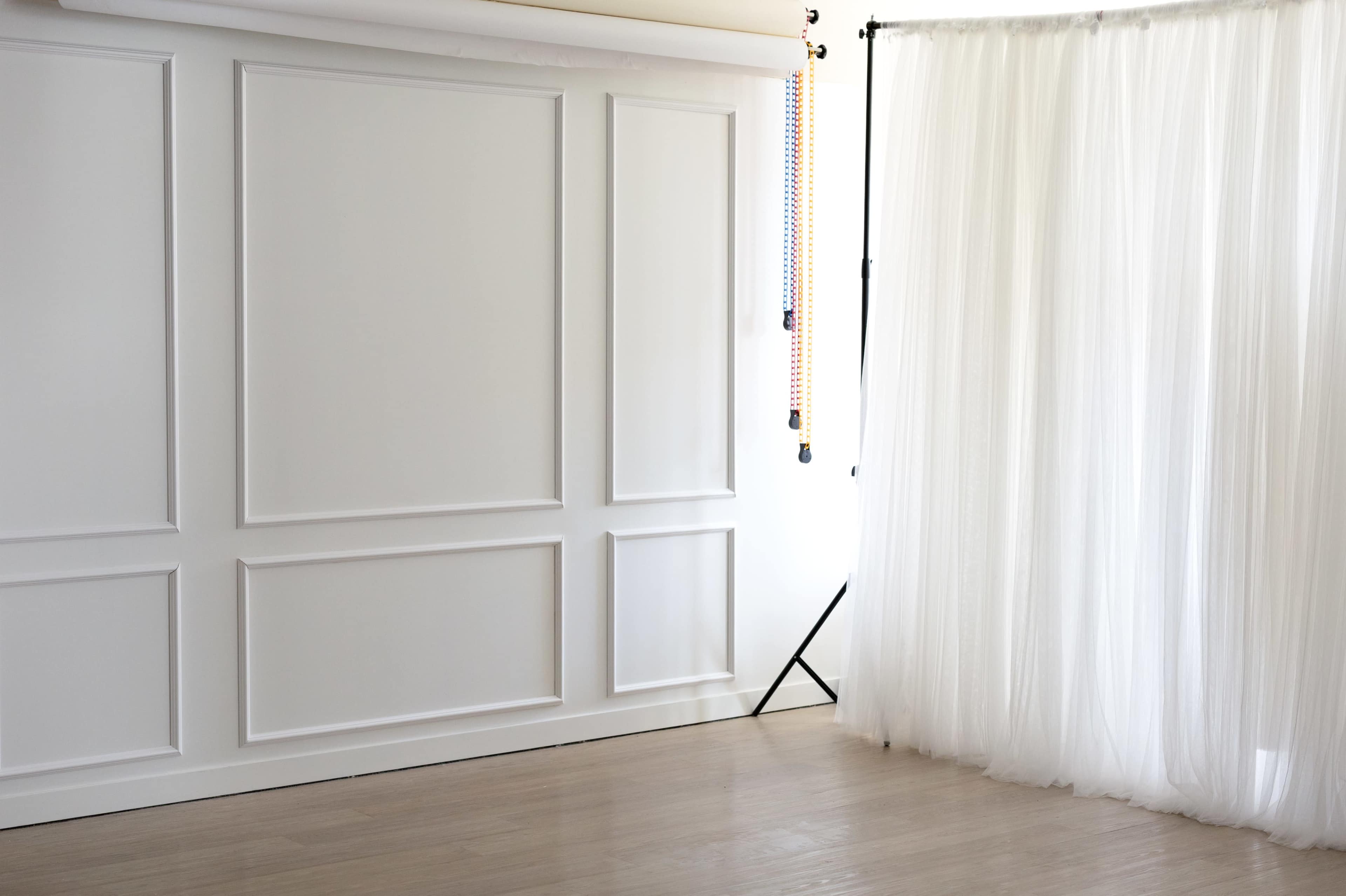 The image shows a minimalist room with a white panelled wall on one side and sheer white curtains illuminated by natural light on the other.