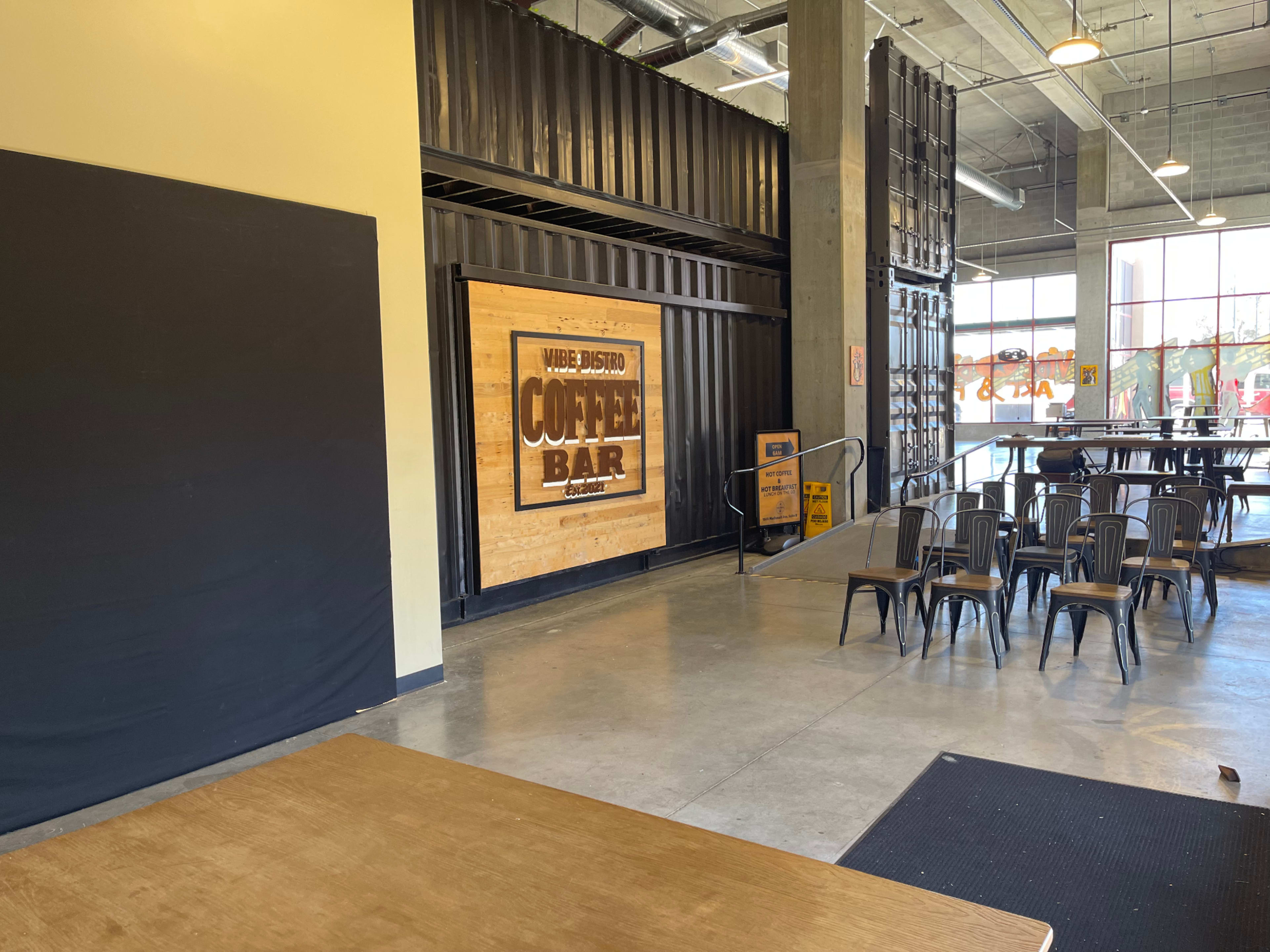 The image shows an interior of a coffee bar featuring a large wooden sign and metal chairs arranged in a spacious, industrial-style setting.