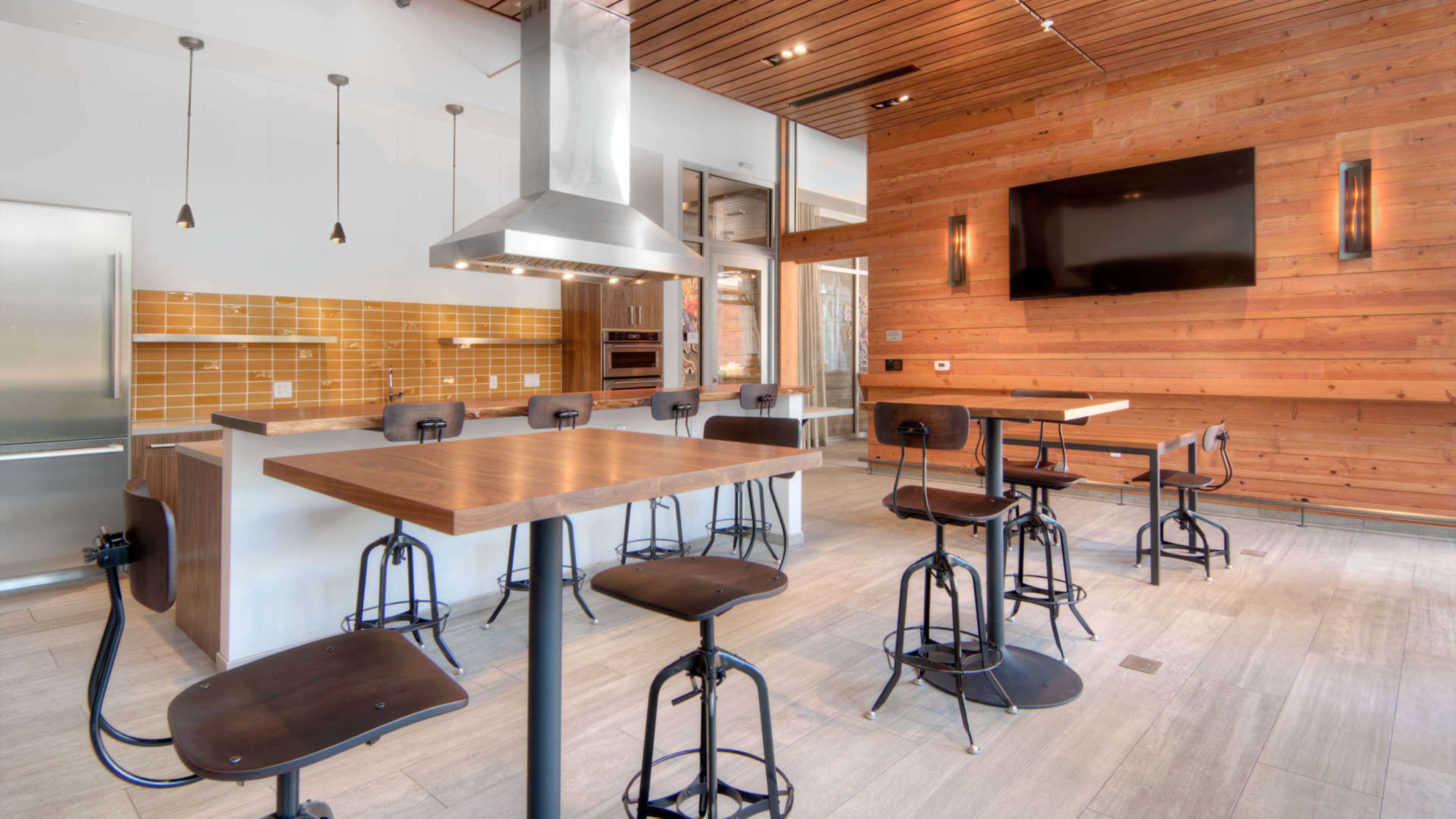 A modern kitchen with stainless steel appliances, wooden walls, and high-top tables with black stools.