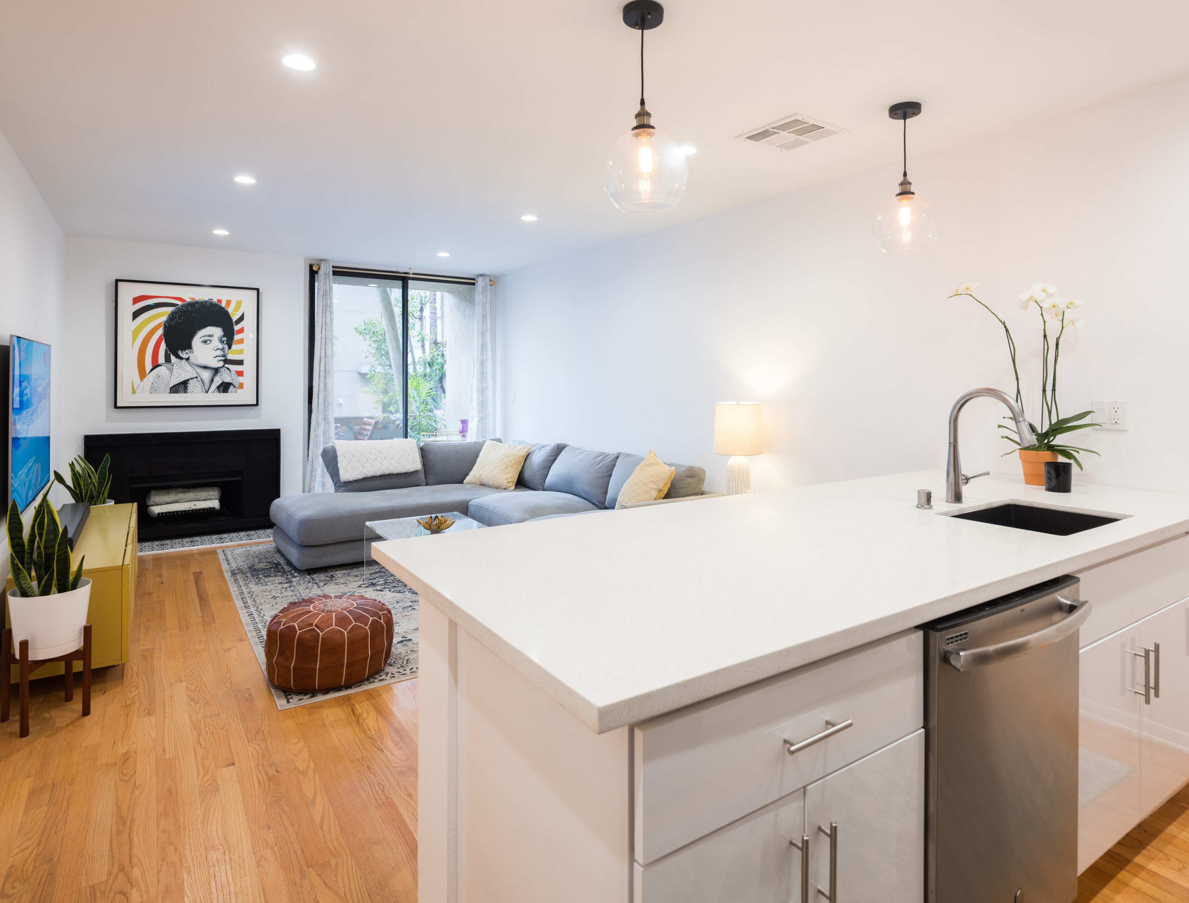 A modern, open-concept living space features a gray sectional sofa, a flat-screen TV, and a kitchen island with a sink and stainless steel dishwasher, illuminated by pendant lights.