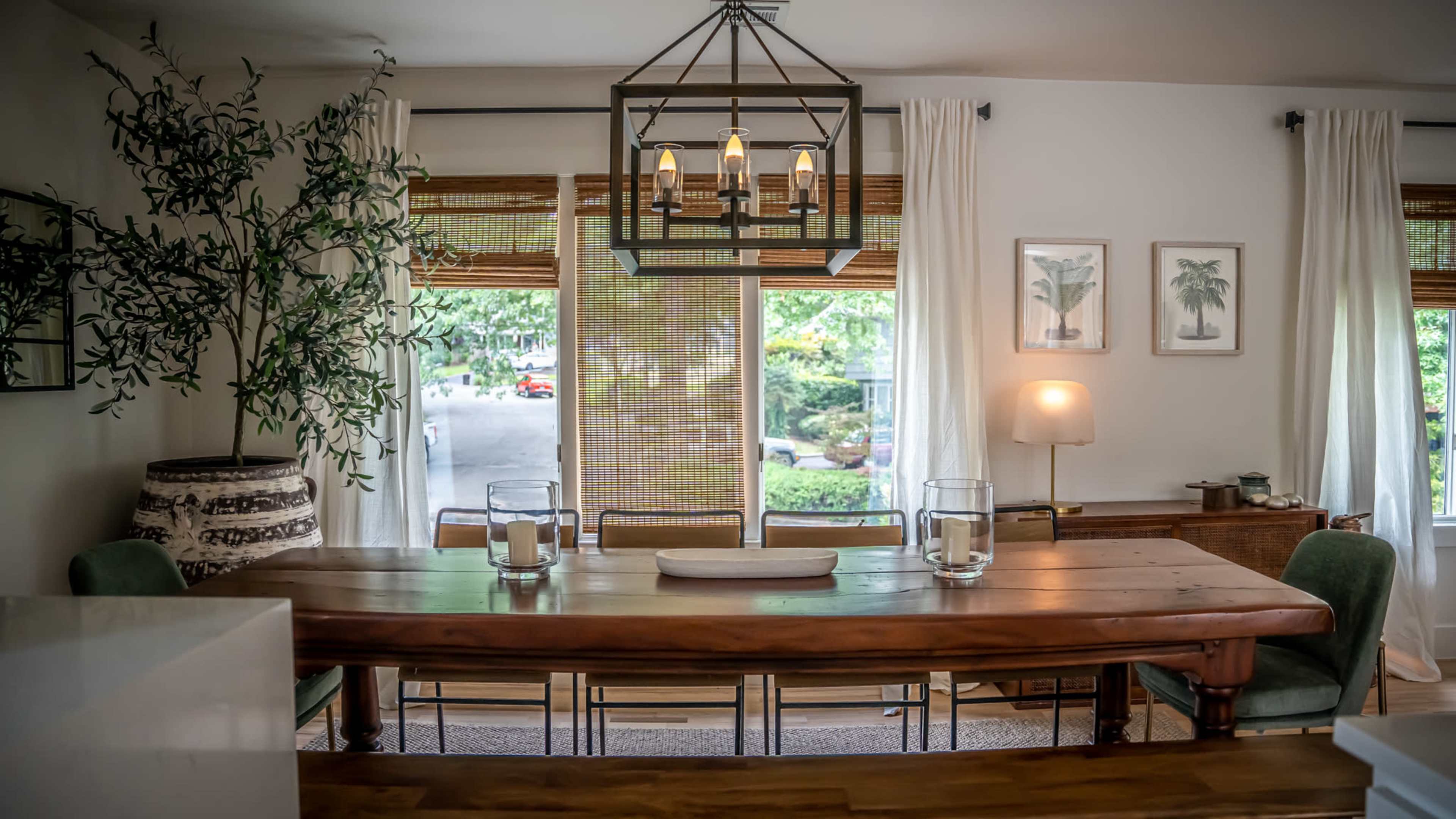 A wooden dining table is set in a bright room with large windows, green chairs, and decorative plants.