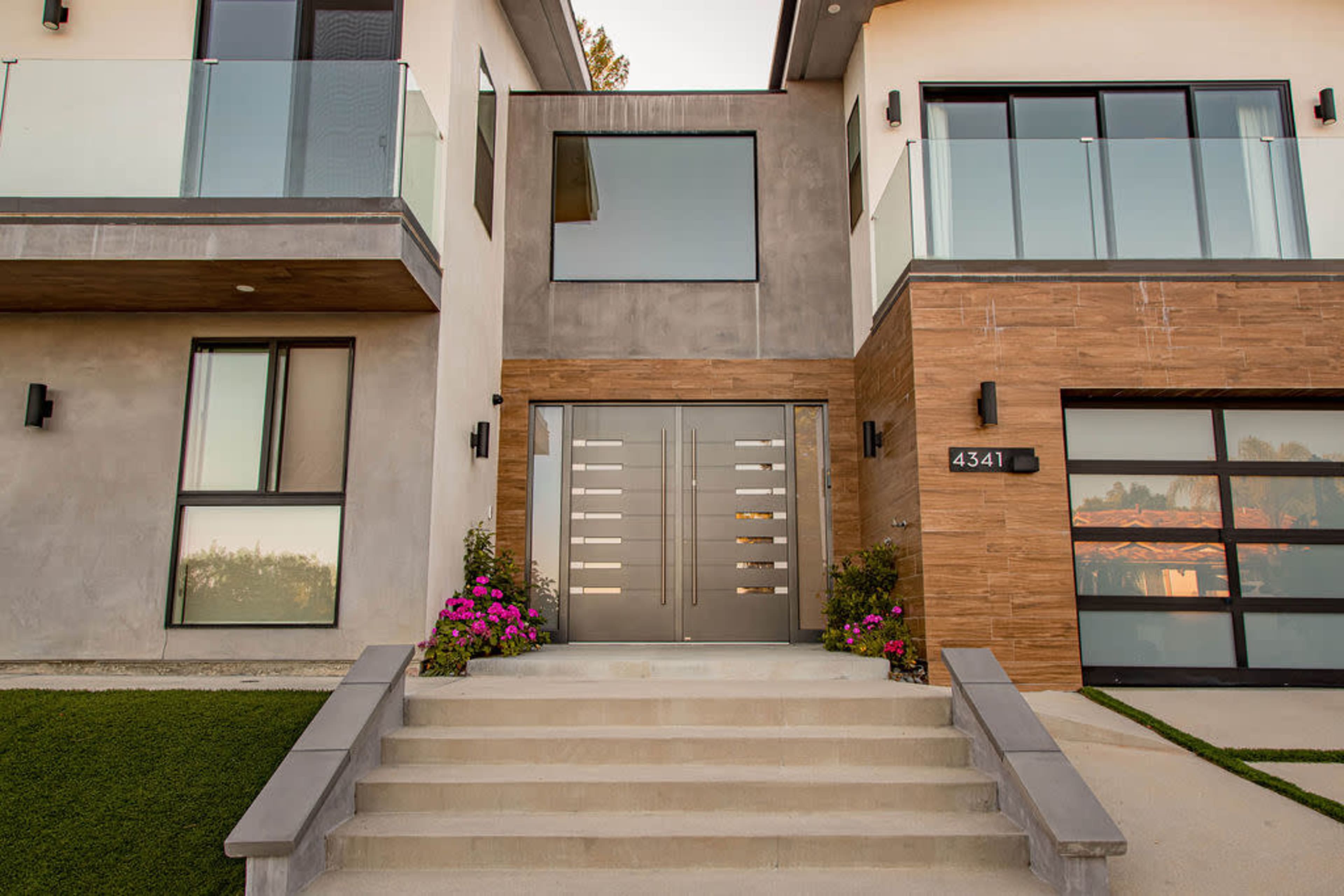 A modern home entrance with a large, double-door entryway flanked by glass railings and landscaped steps leading to the door.