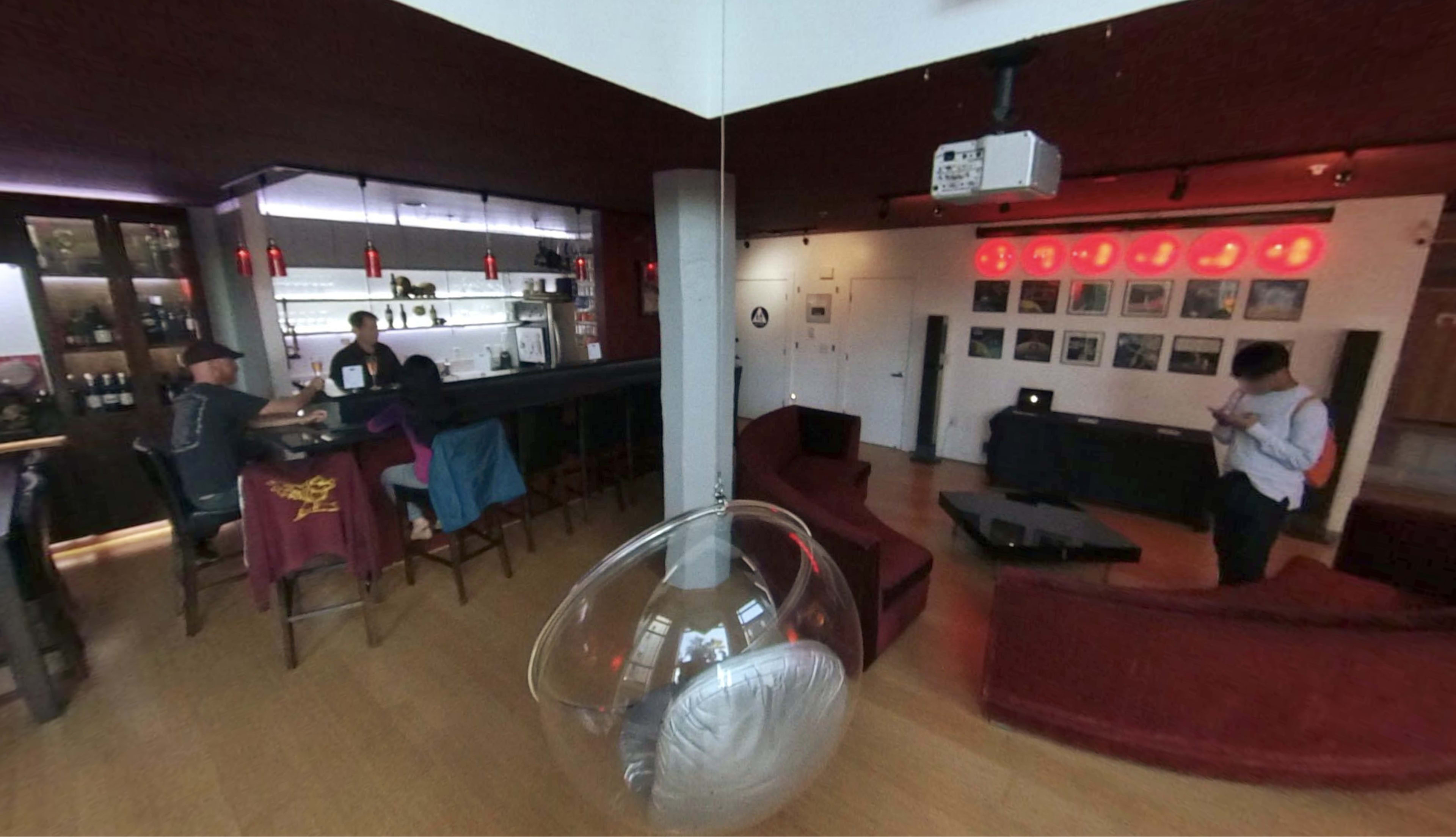 A modern bar area with a curved counter, seating along the bar, red accents, and a person standing near a wall lined with framed pictures.