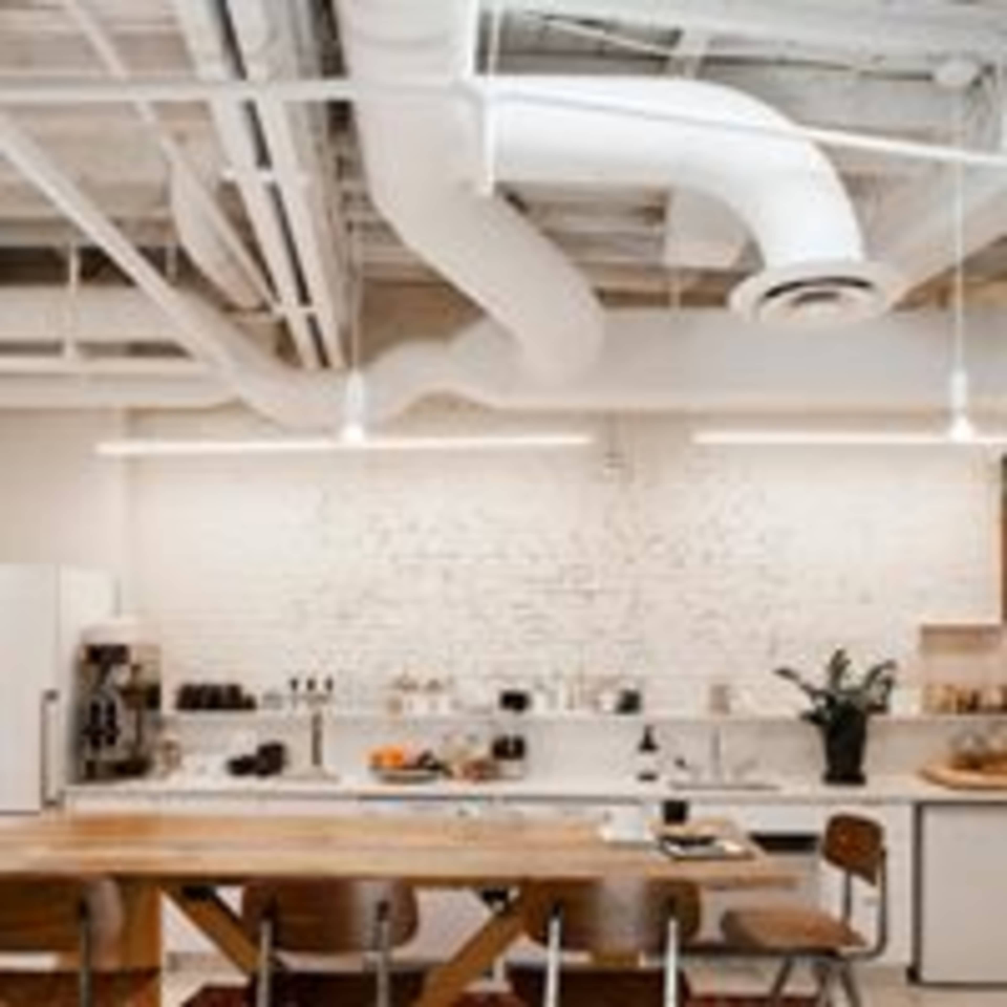 The image depicts a modern kitchen with a large wooden table, shelves filled with kitchenware, and exposed ductwork overhead.
