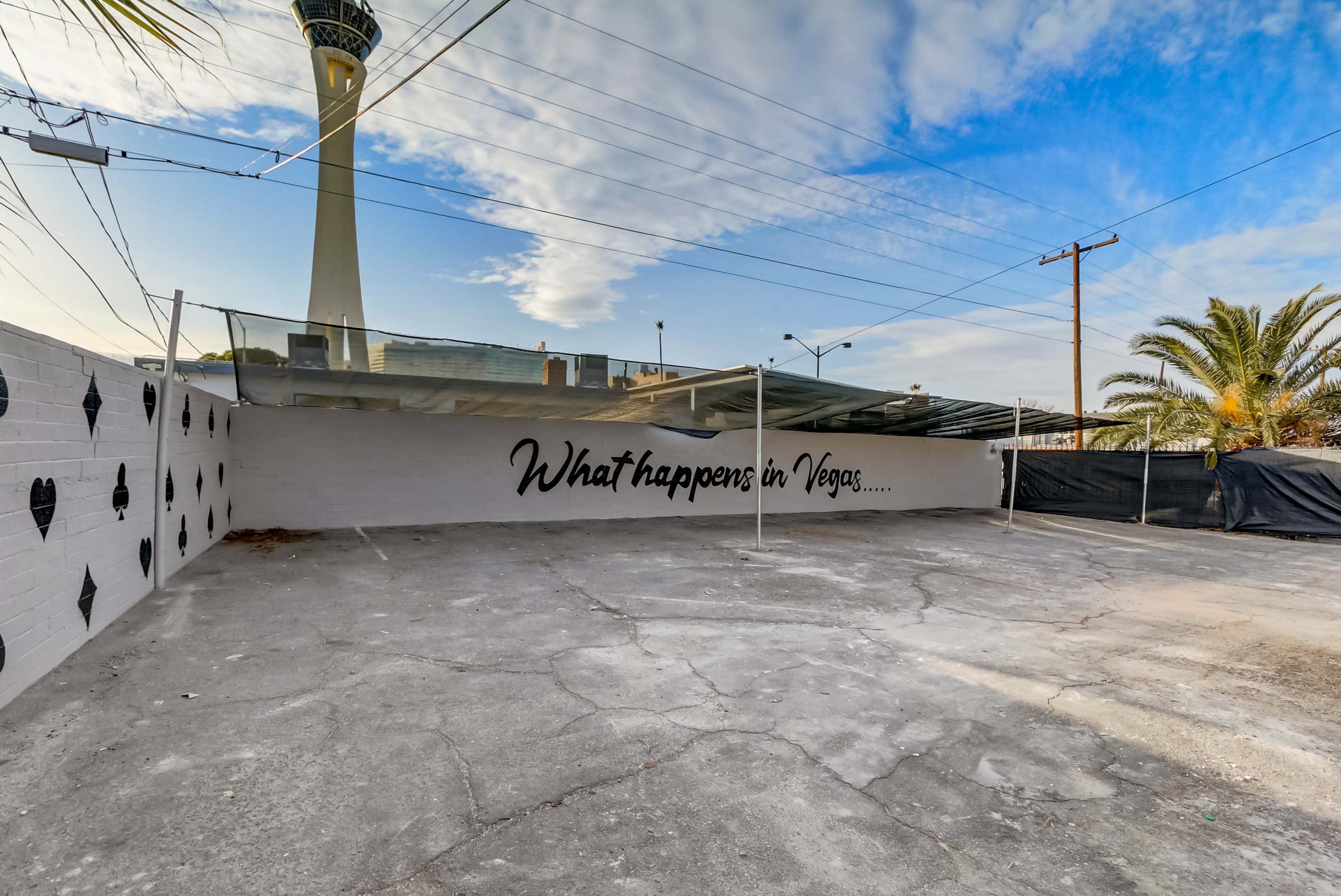 The image shows a vacant parking lot with a mural that reads "What happens in Vegas..." and features the Stratosphere tower in the background.