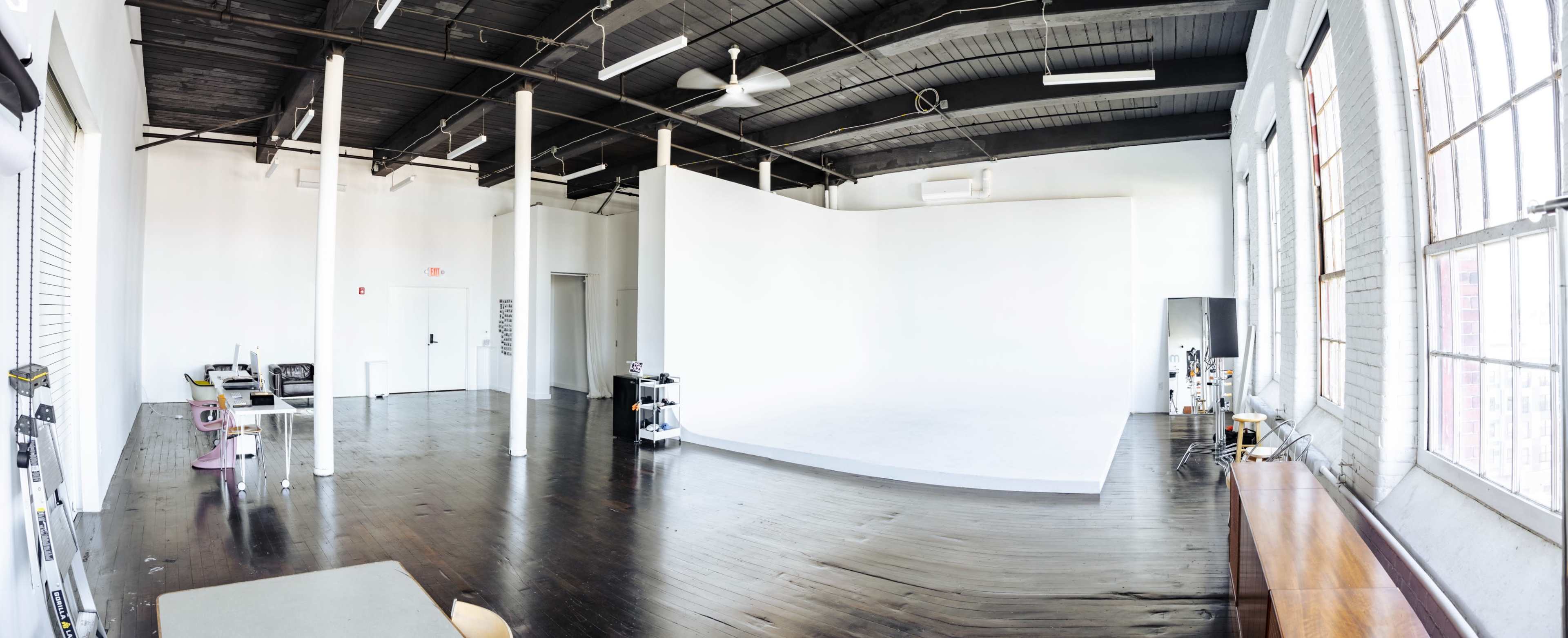 A spacious studio with wooden floors, white walls, and large windows, featuring a backdrop area and minimal furnishings.
