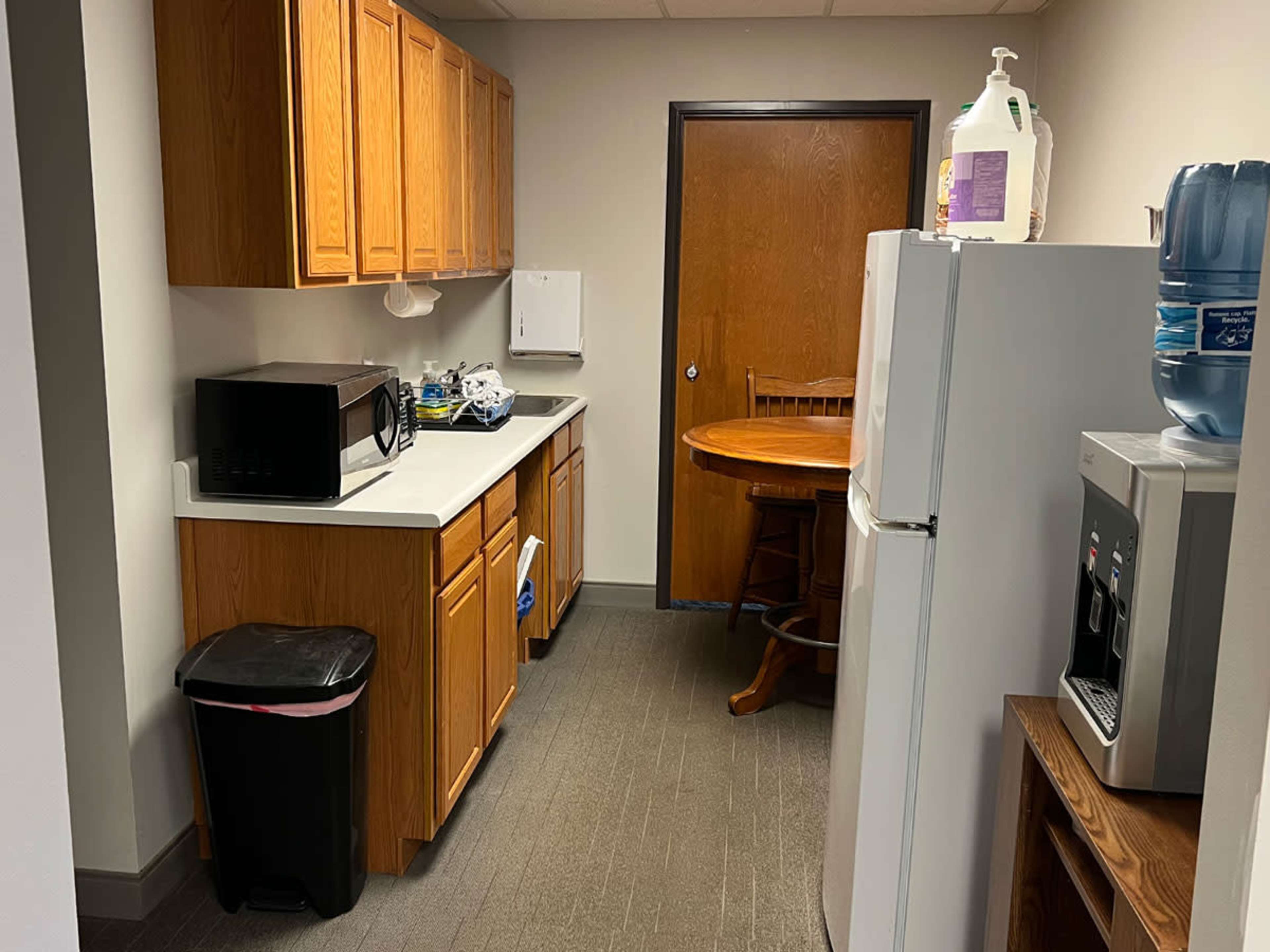 A small kitchen area with wooden cabinets, a microwave, a round wooden table, and a refrigerator beside a water cooler.