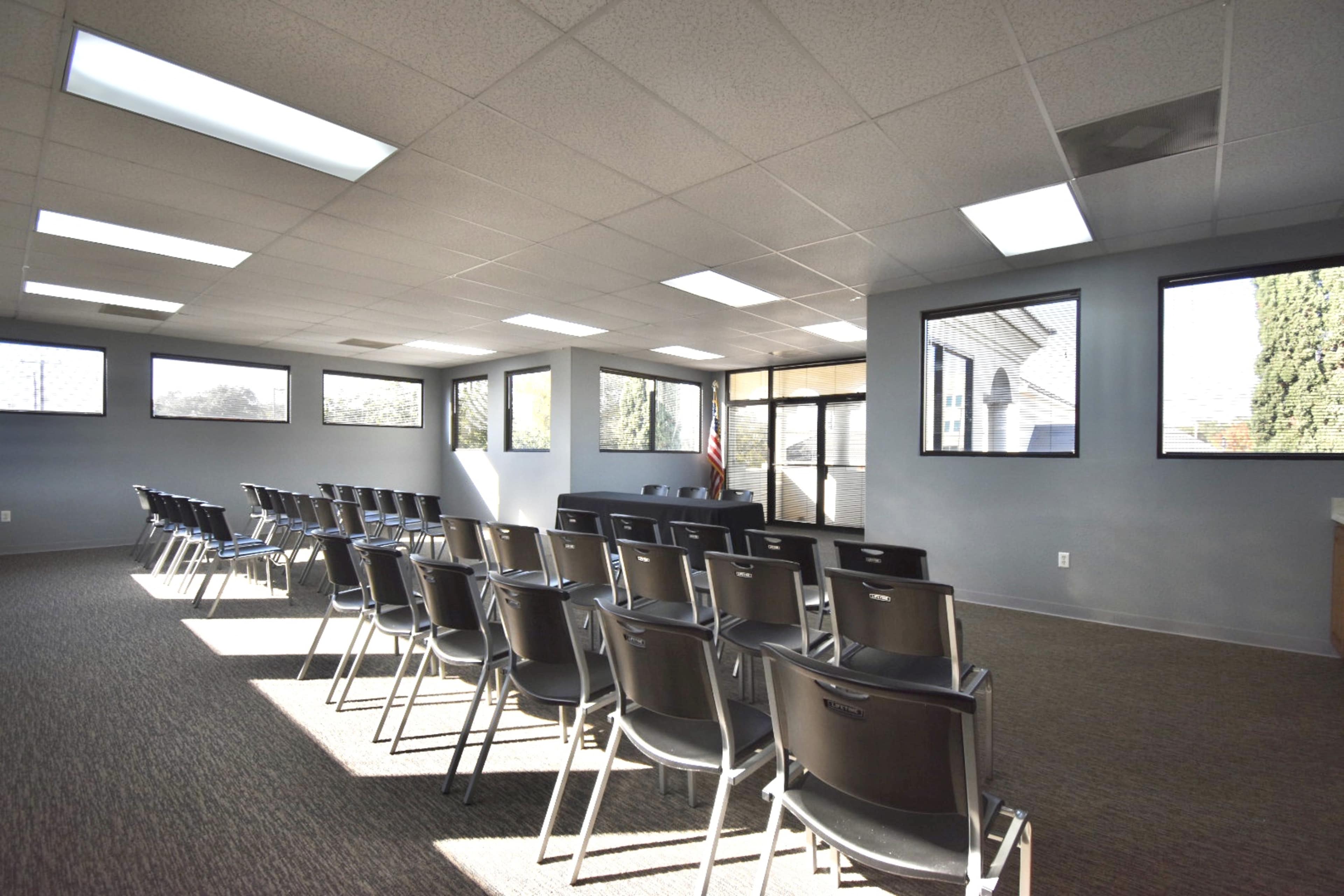 A vacant meeting room features rows of black chairs facing a front area with large windows and a door leading outside.