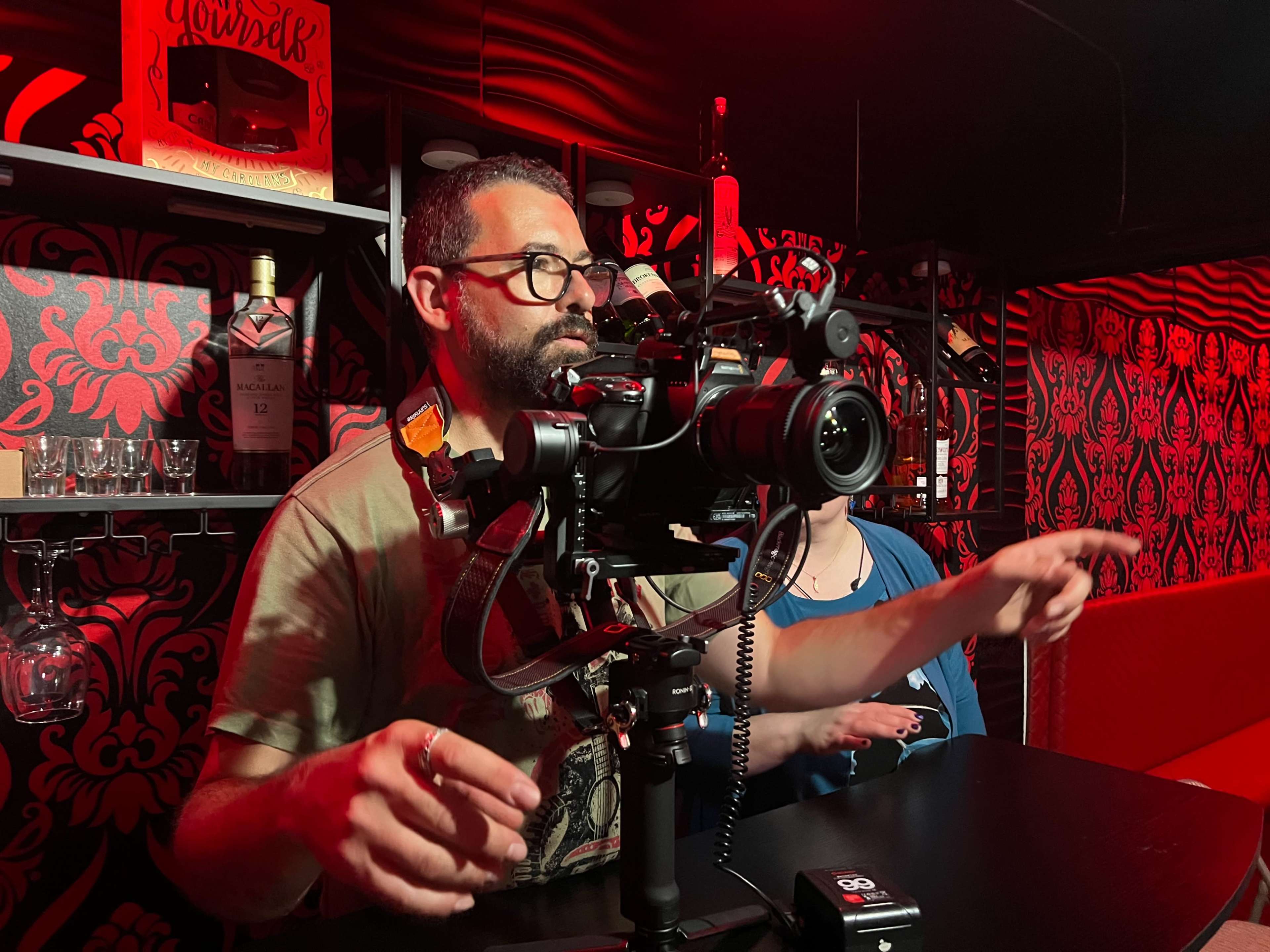 A man operates a camera on a stabilizer in a dimly lit room with patterned red walls and shelves displaying bottles and glasses.