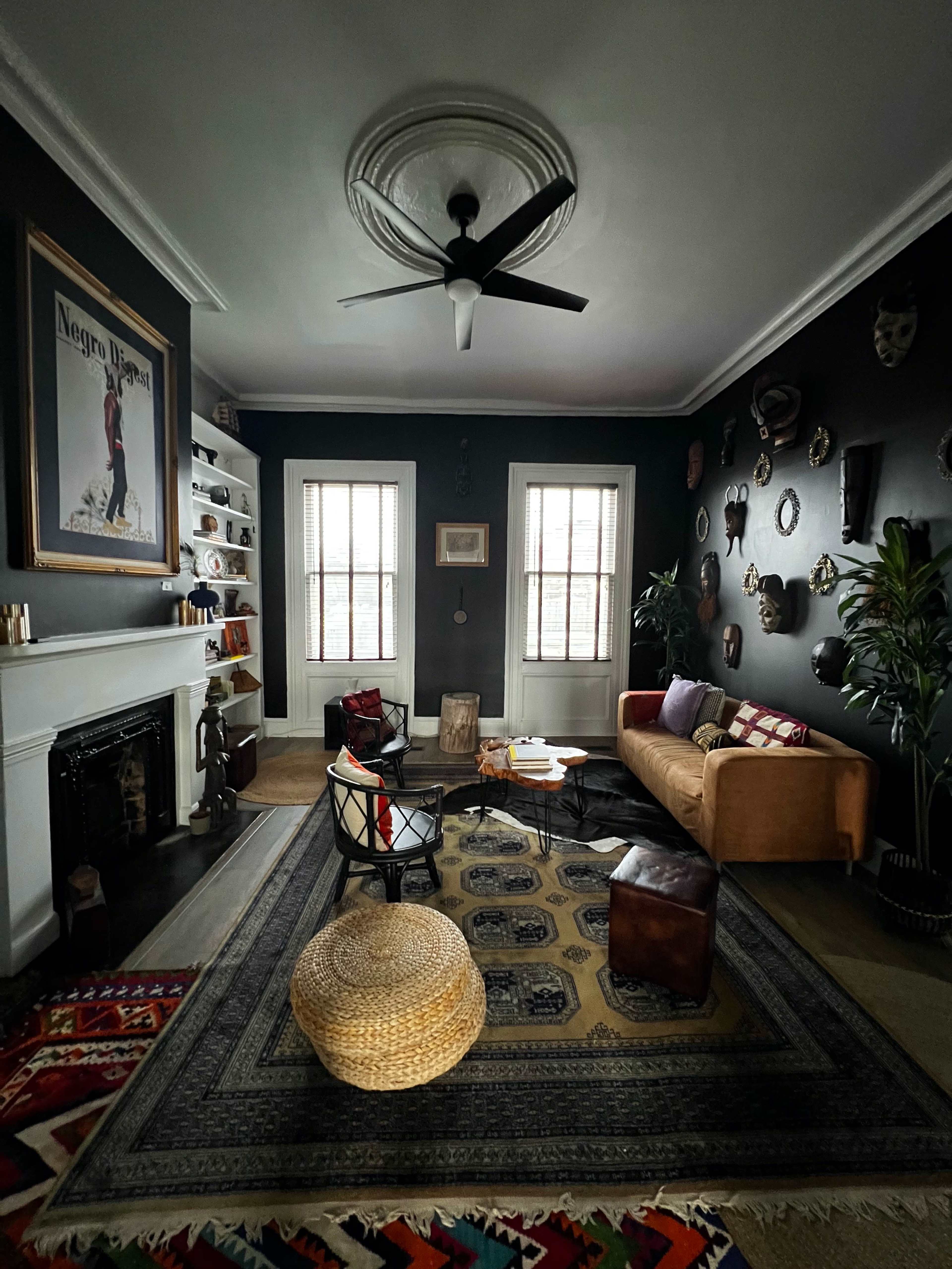 A dark-colored living room featuring a mix of modern and vintage furnishings, including a ceiling fan, a fireplace, a large wall poster, and various decorative masks on the walls.