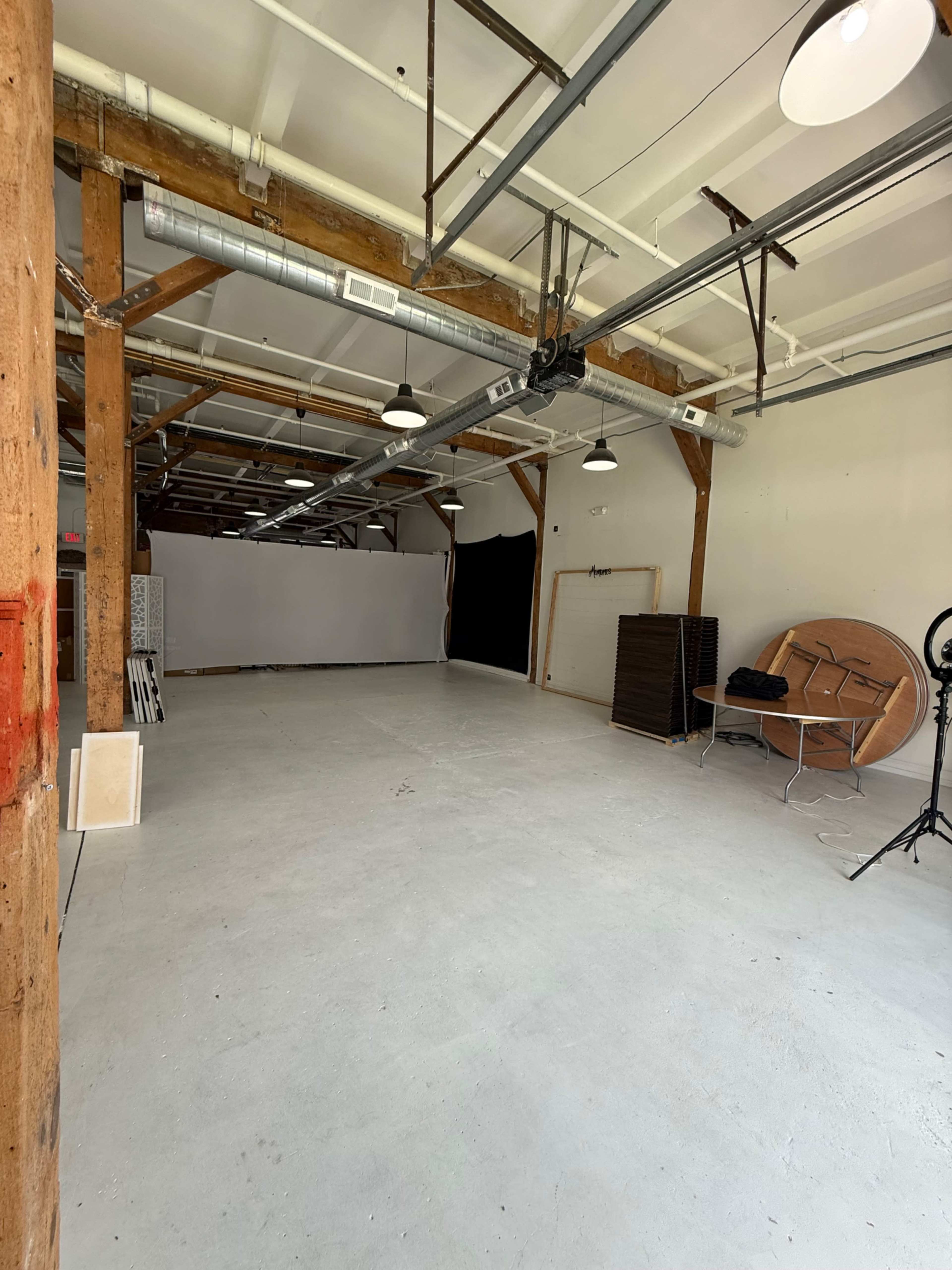 The image shows a spacious interior of a vacant studio with wooden beams, overhead lighting, and minimal furnishings.