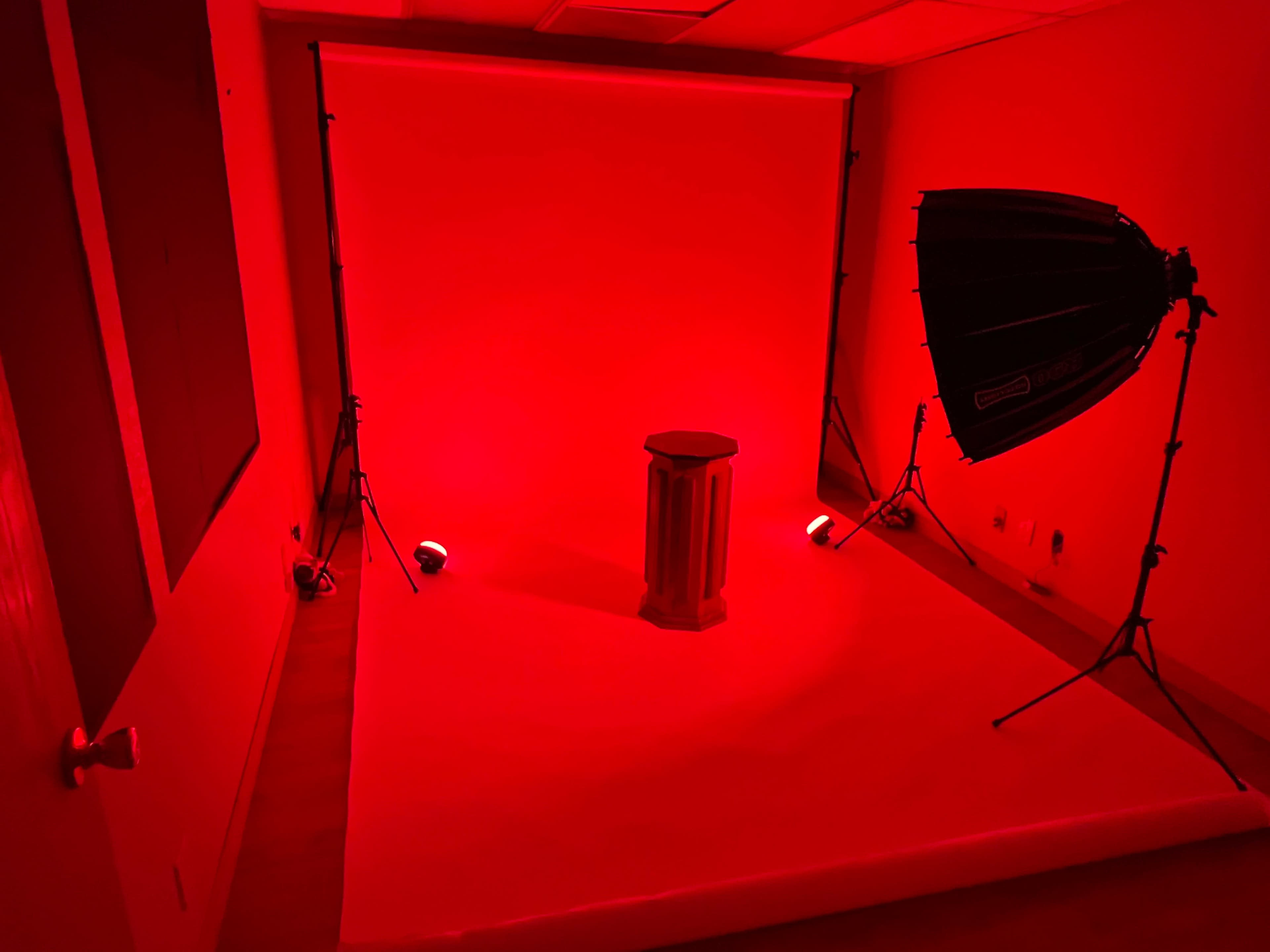 A photography set with a red backdrop, spotlighting a pedestal in the center, accompanied by lighting equipment on the side.