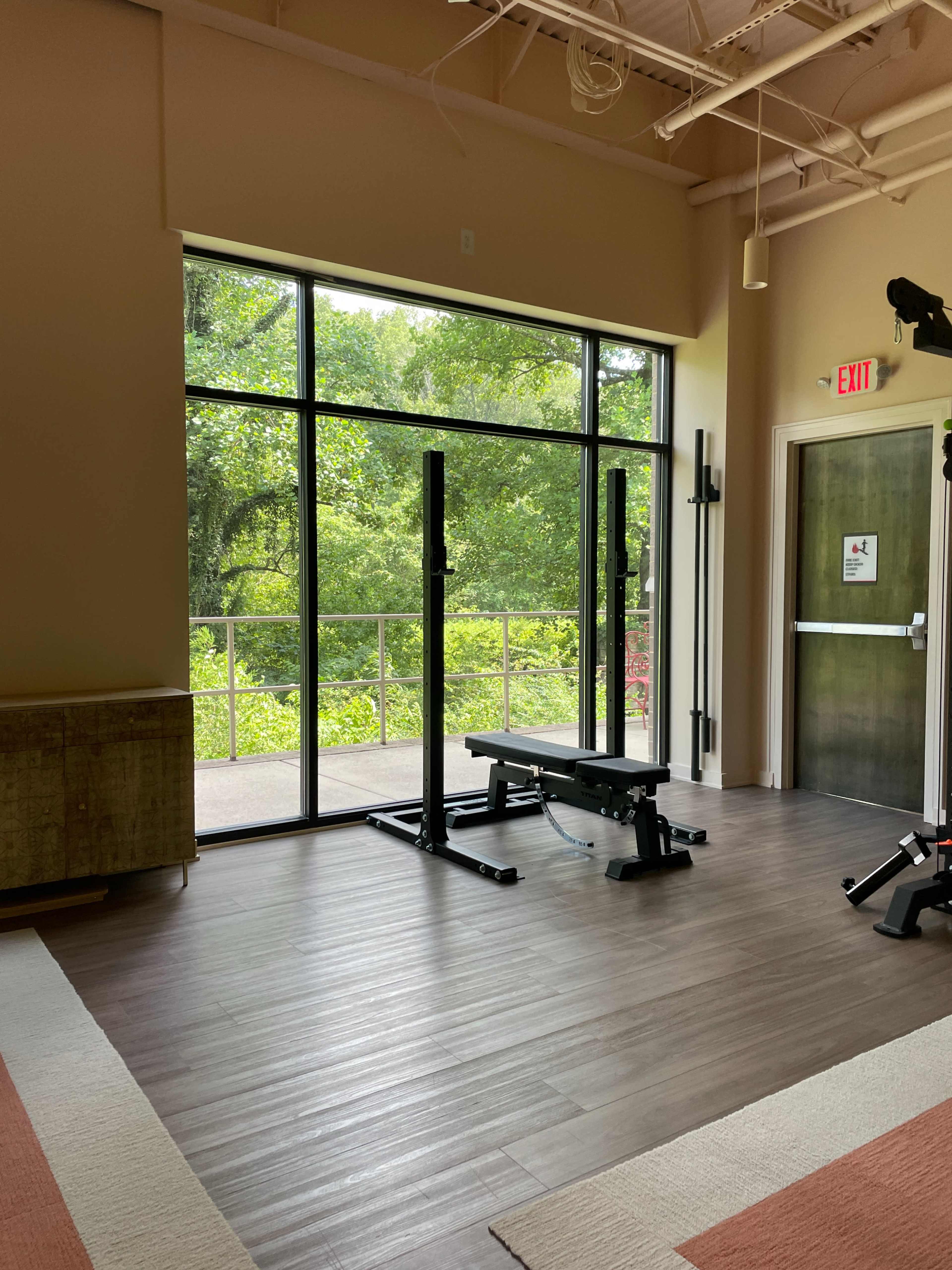 The image shows a gym area with a weight bench and squat rack positioned near large windows overlooking a green outdoor landscape.