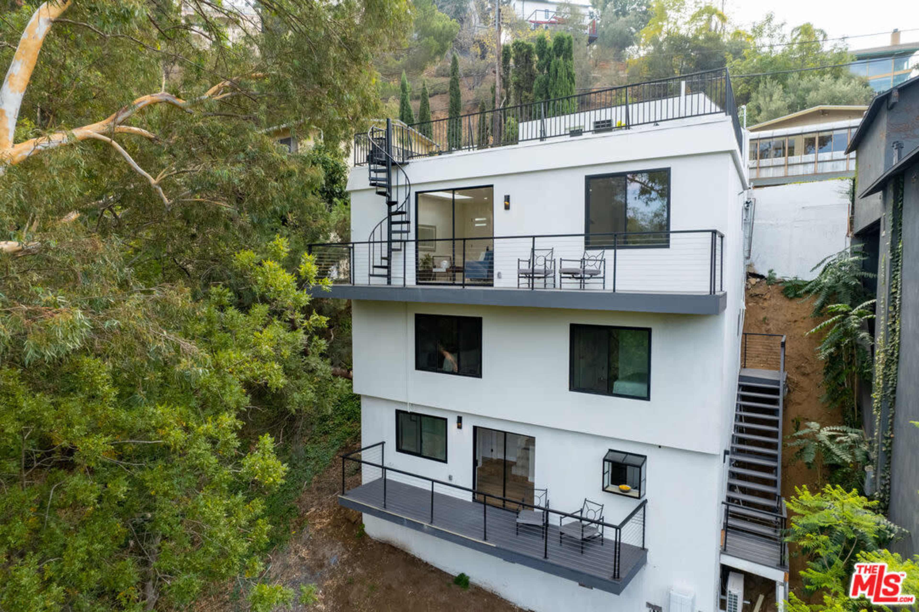 The image shows a three-story modern white house with large windows, a balcony, and a staircase, set on a hillside surrounded by trees.