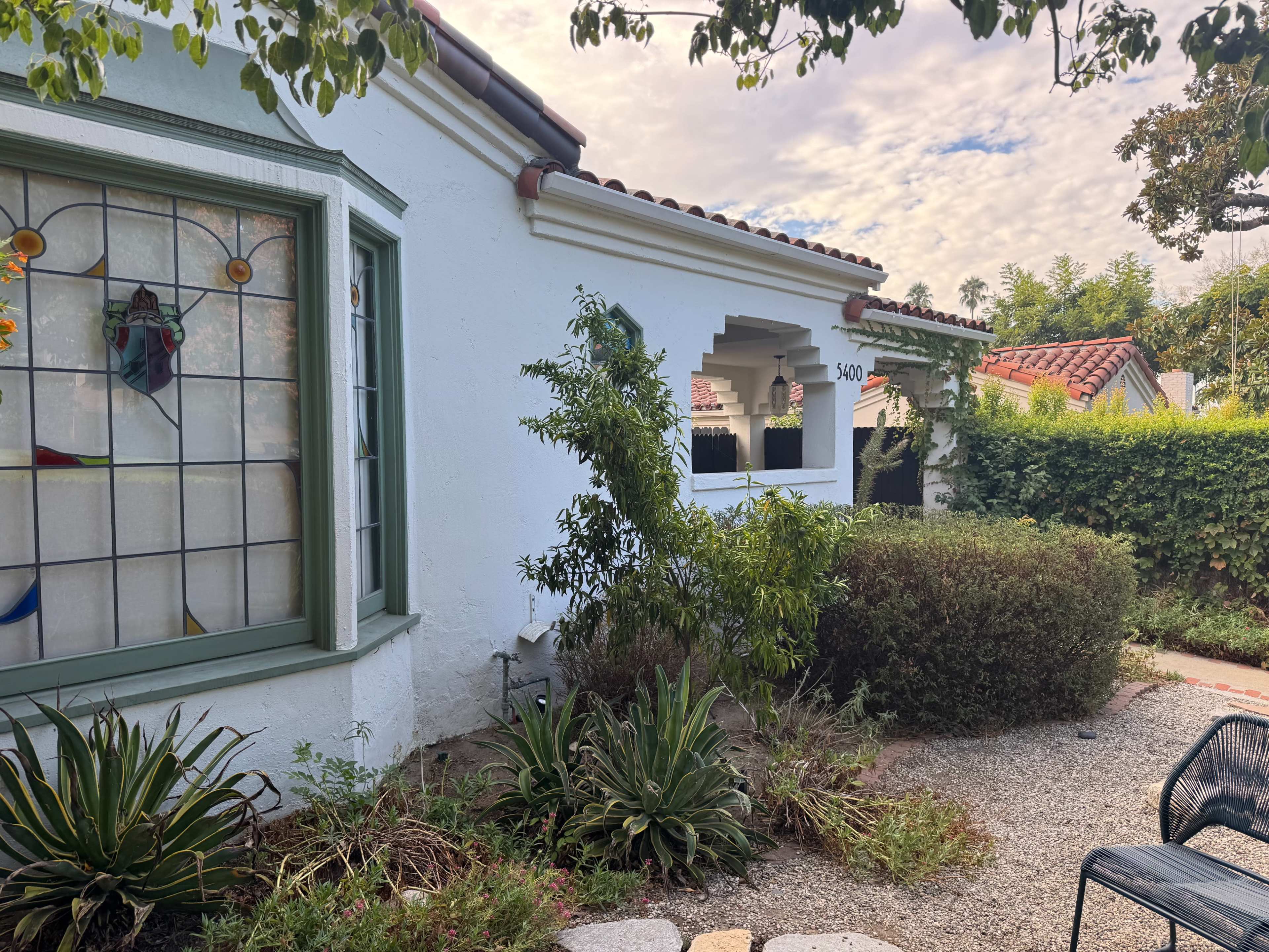 The image shows a white stucco house with a green-trimmed window and a landscaped garden featuring various plants and gravel pathways.