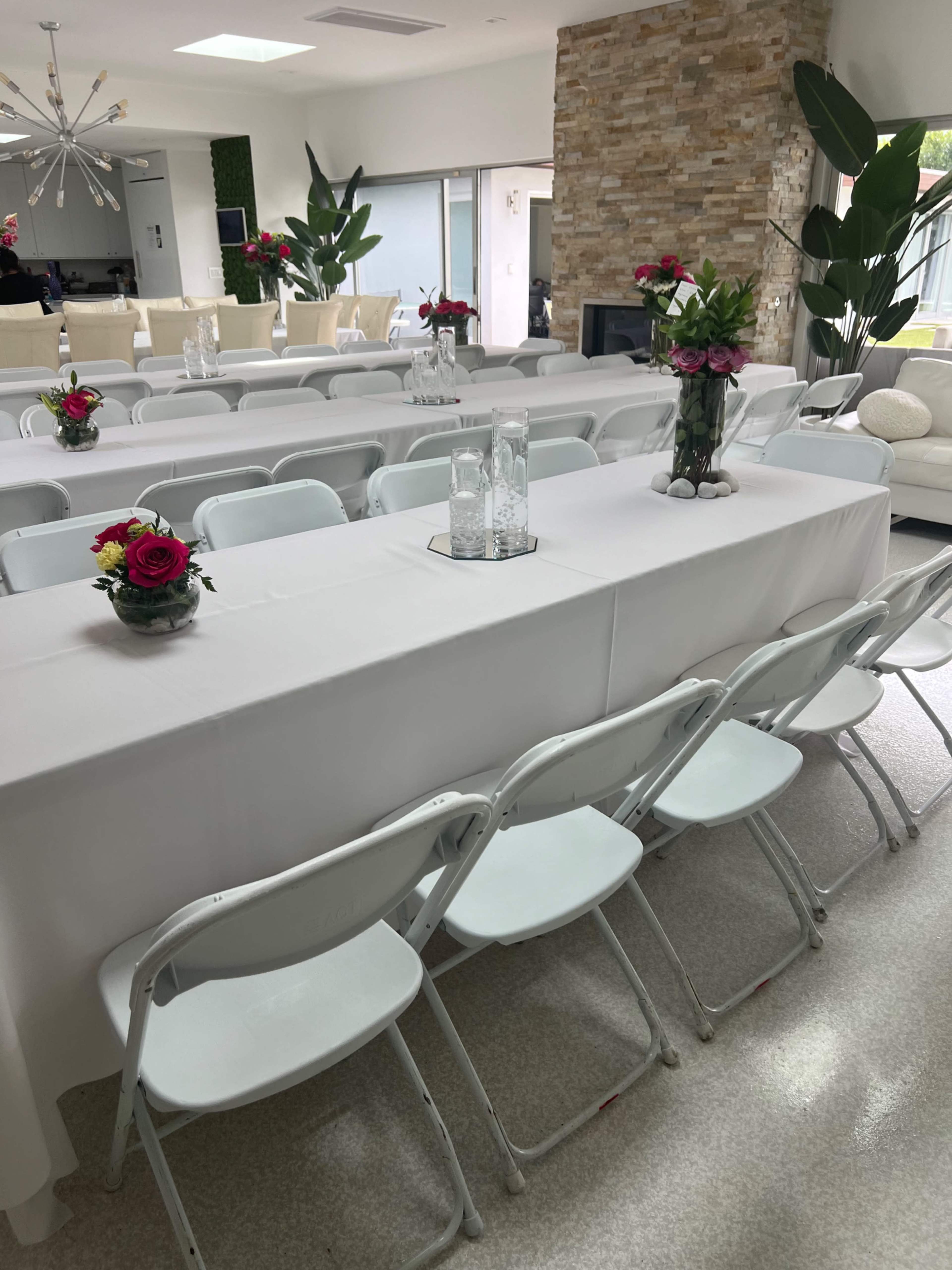 A spacious, elegantly arranged indoor event space features white tables with decorative centerpieces and surrounding folding chairs.
