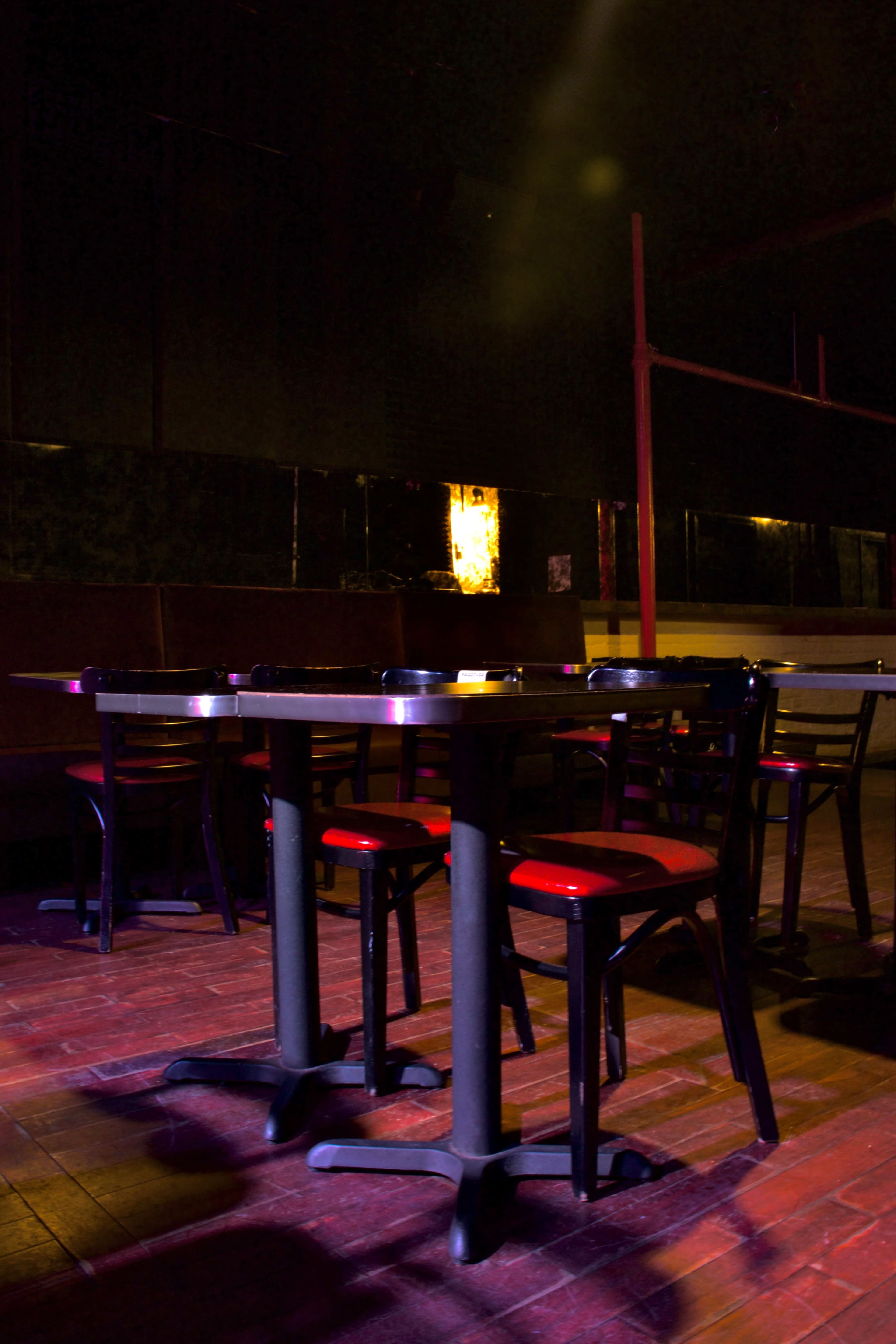 A dimly lit room features tables and chairs with red padding arranged on a wooden floor.