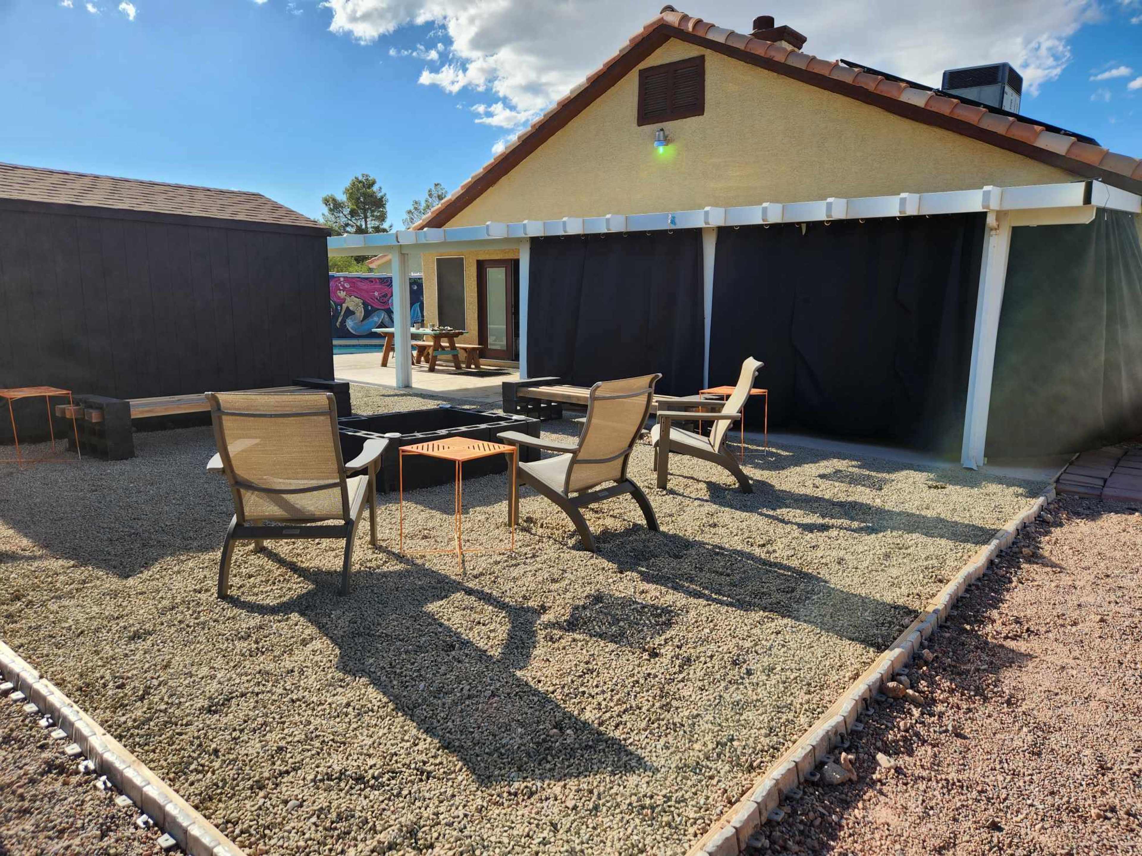 A yard features gravel landscaping with two lounge chairs and a small table near a fire pit, beside a yellow house with a black wall.