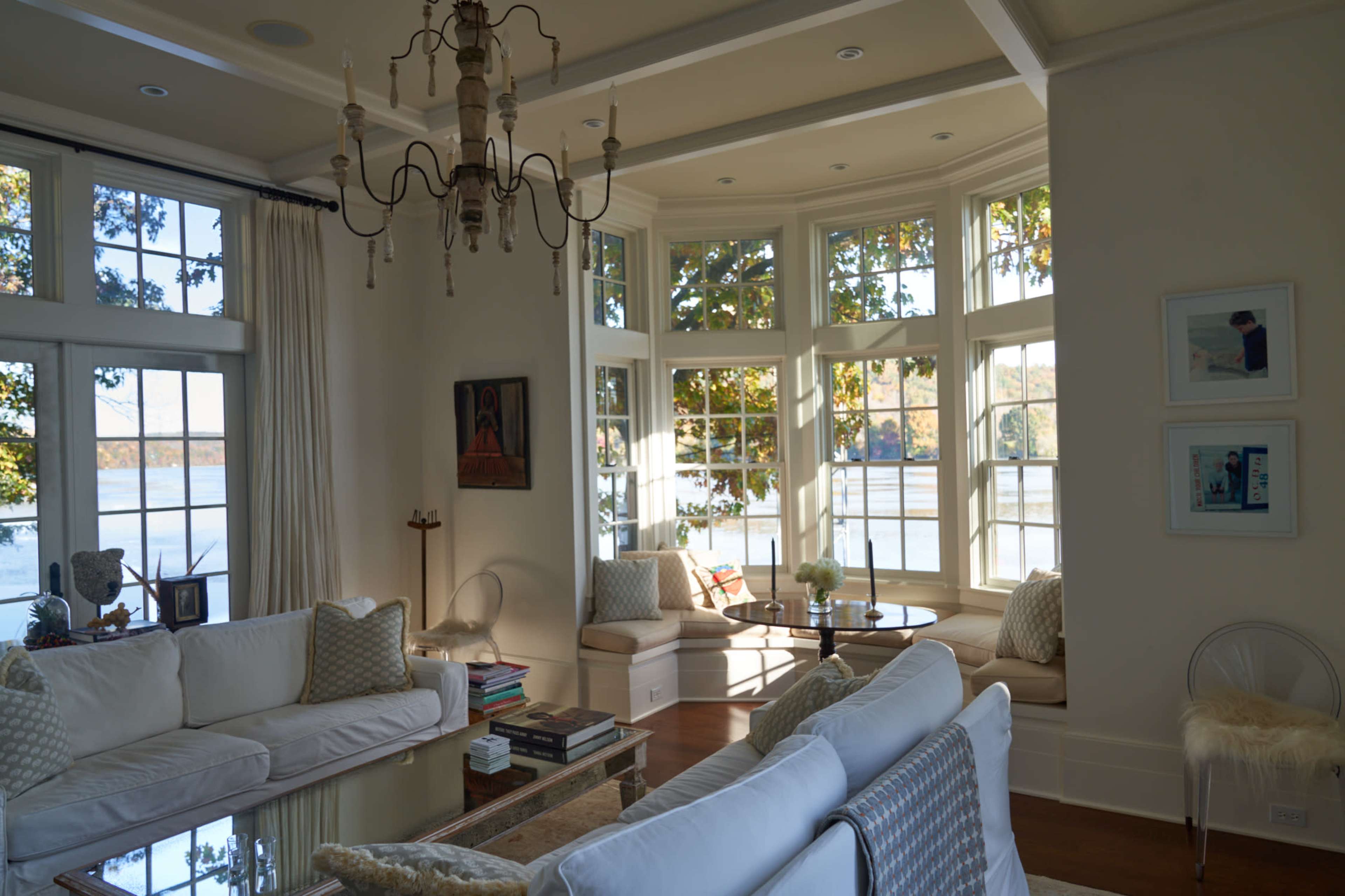 A bright living room features large windows overlooking water, with a chandelier and white sofas arranged around a coffee table.