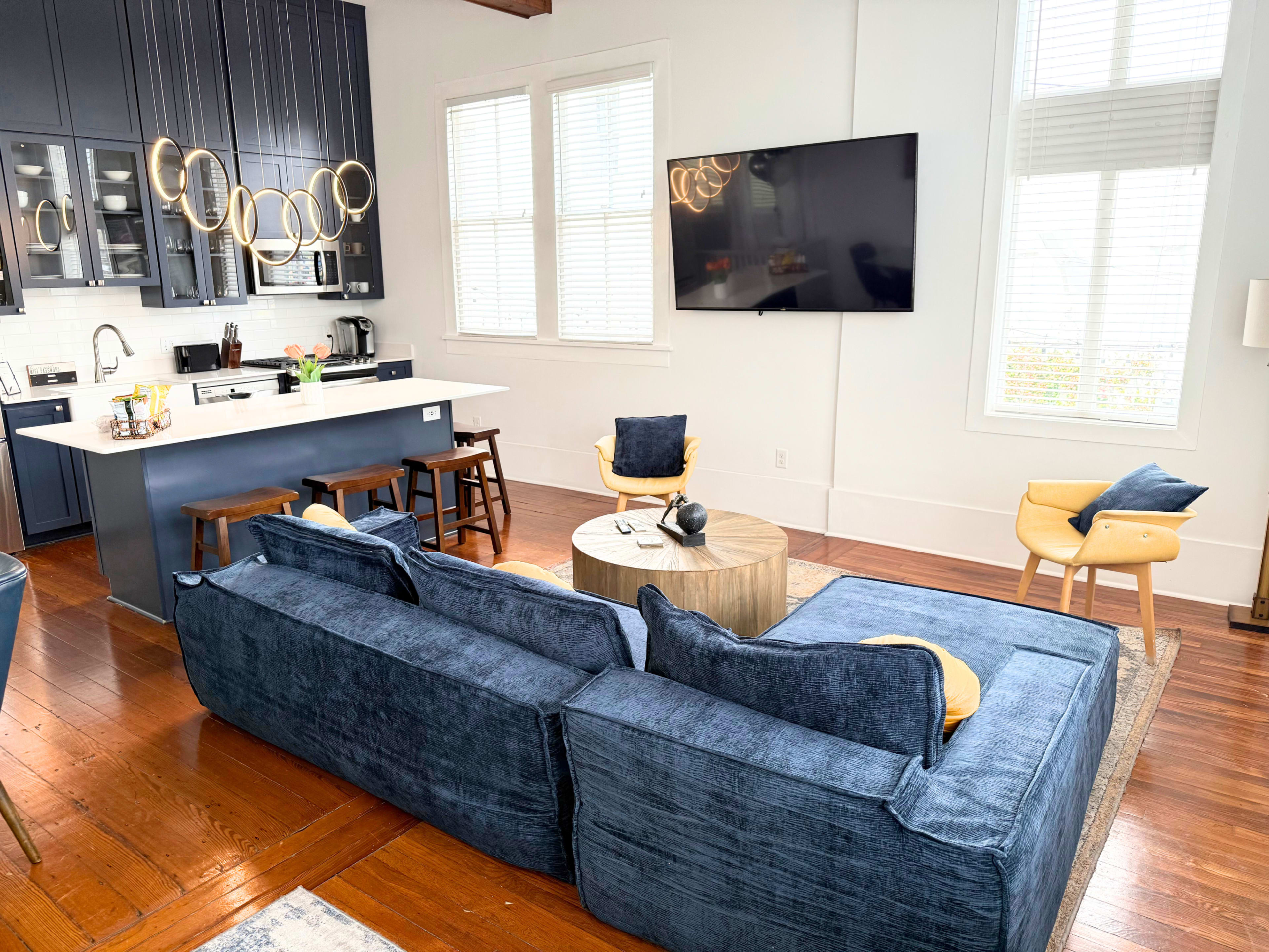 The image shows a modern living space featuring a blue sectional sofa, a round wooden coffee table, and a kitchen area with dark cabinetry and a wall-mounted television.