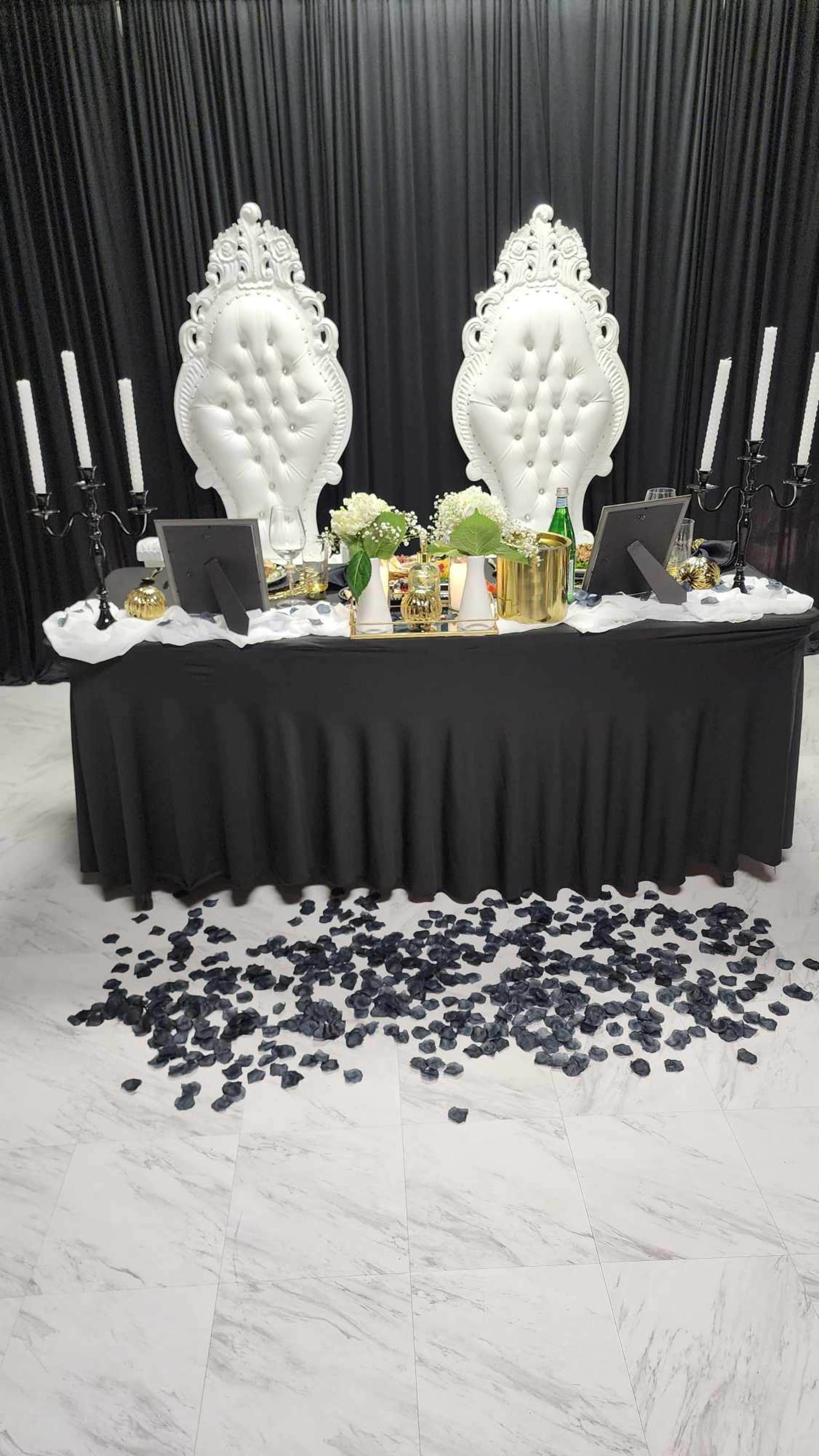 A black-topped table is set with decor and two ornate white chairs against a black backdrop, with black rose petals scattered on the floor.