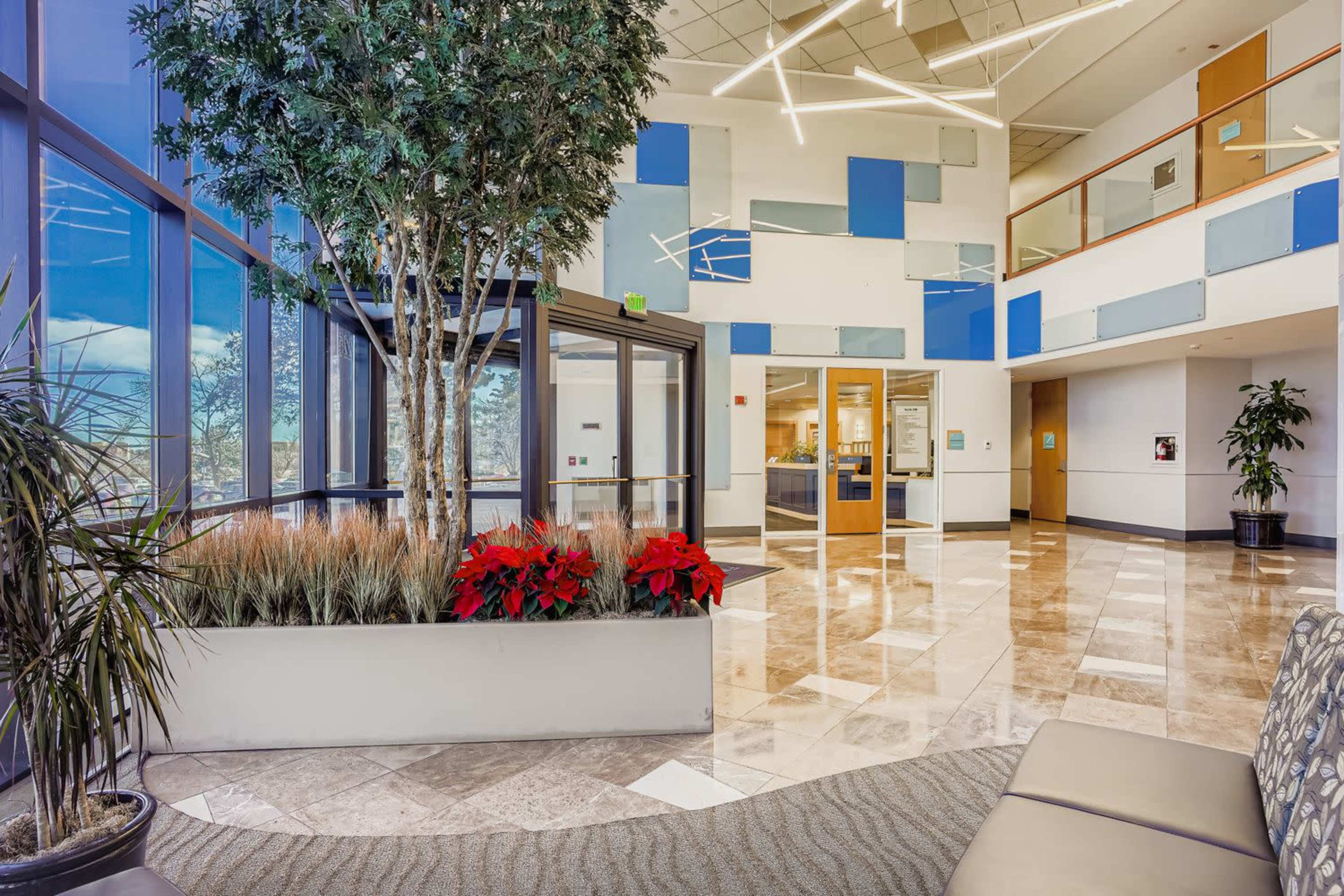 The image shows a modern lobby with large windows, potted plants, and decorative foliage, featuring polished marble flooring and contemporary lighting.