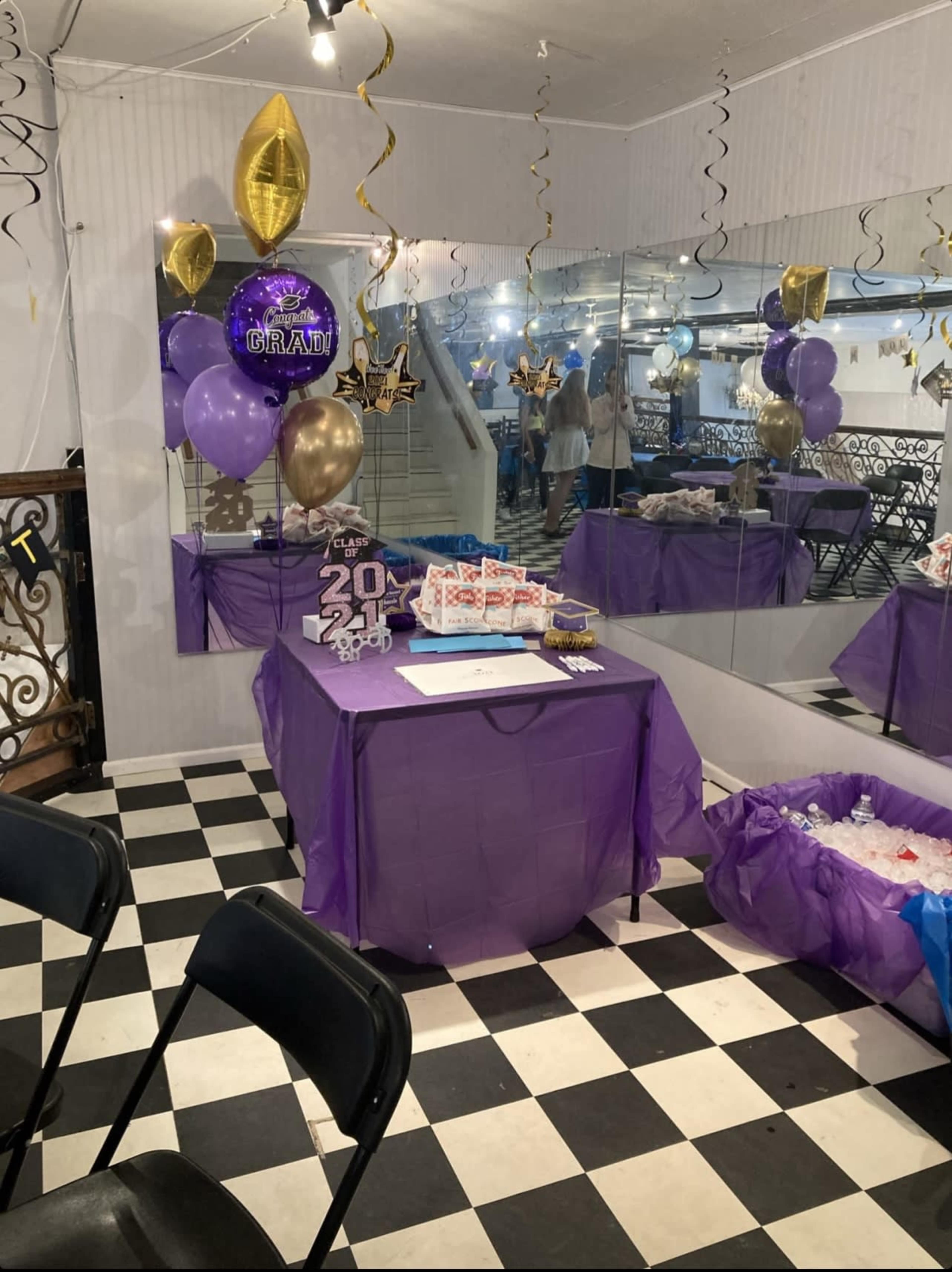 A party setup featuring a purple table with gifts, balloons, and decorations, set against a backdrop of mirrored walls and a black-and-white checkered floor.