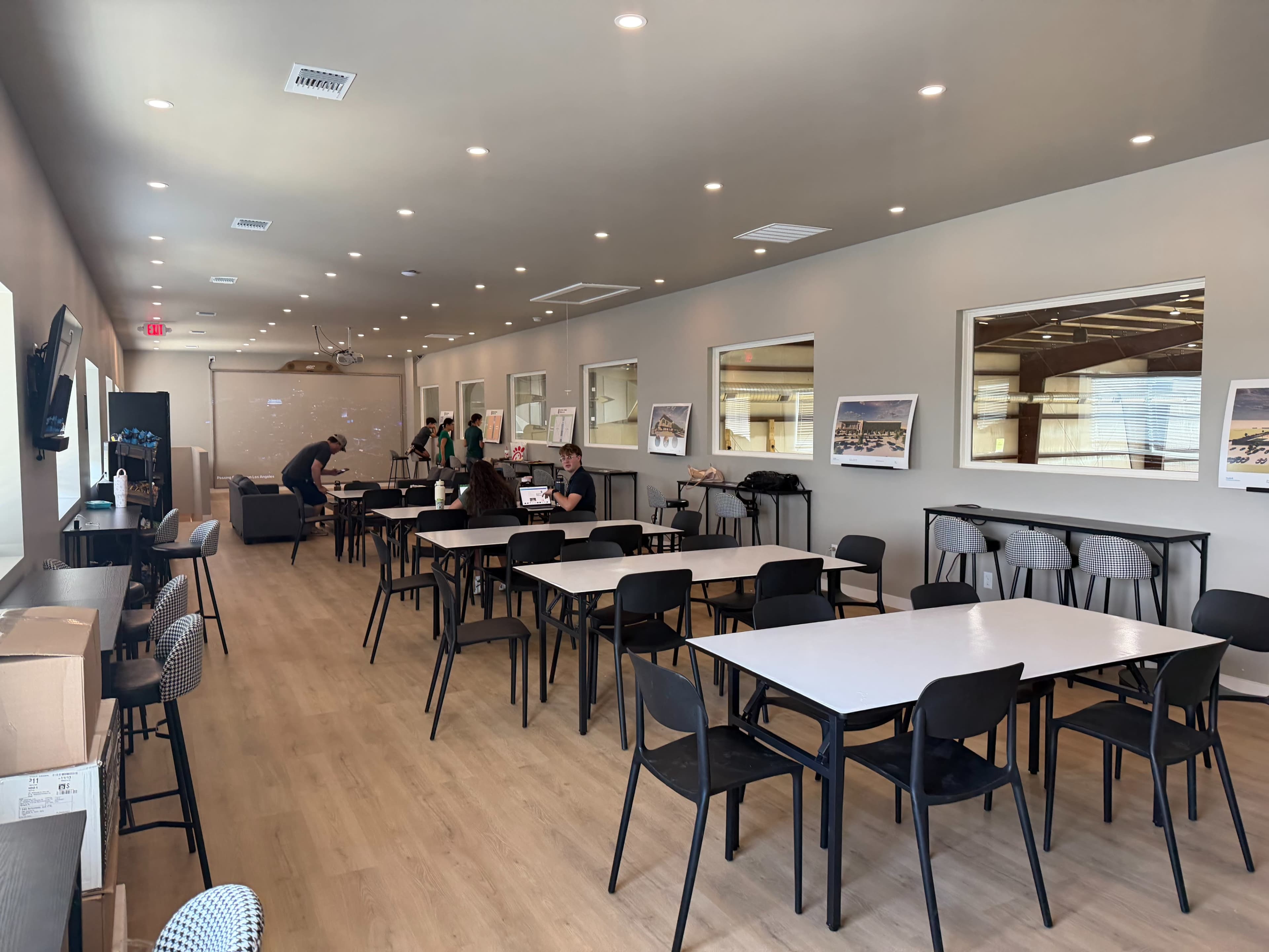 A spacious indoor room features multiple tables and chairs, with a few people engaged in activities and large windows allowing natural light.