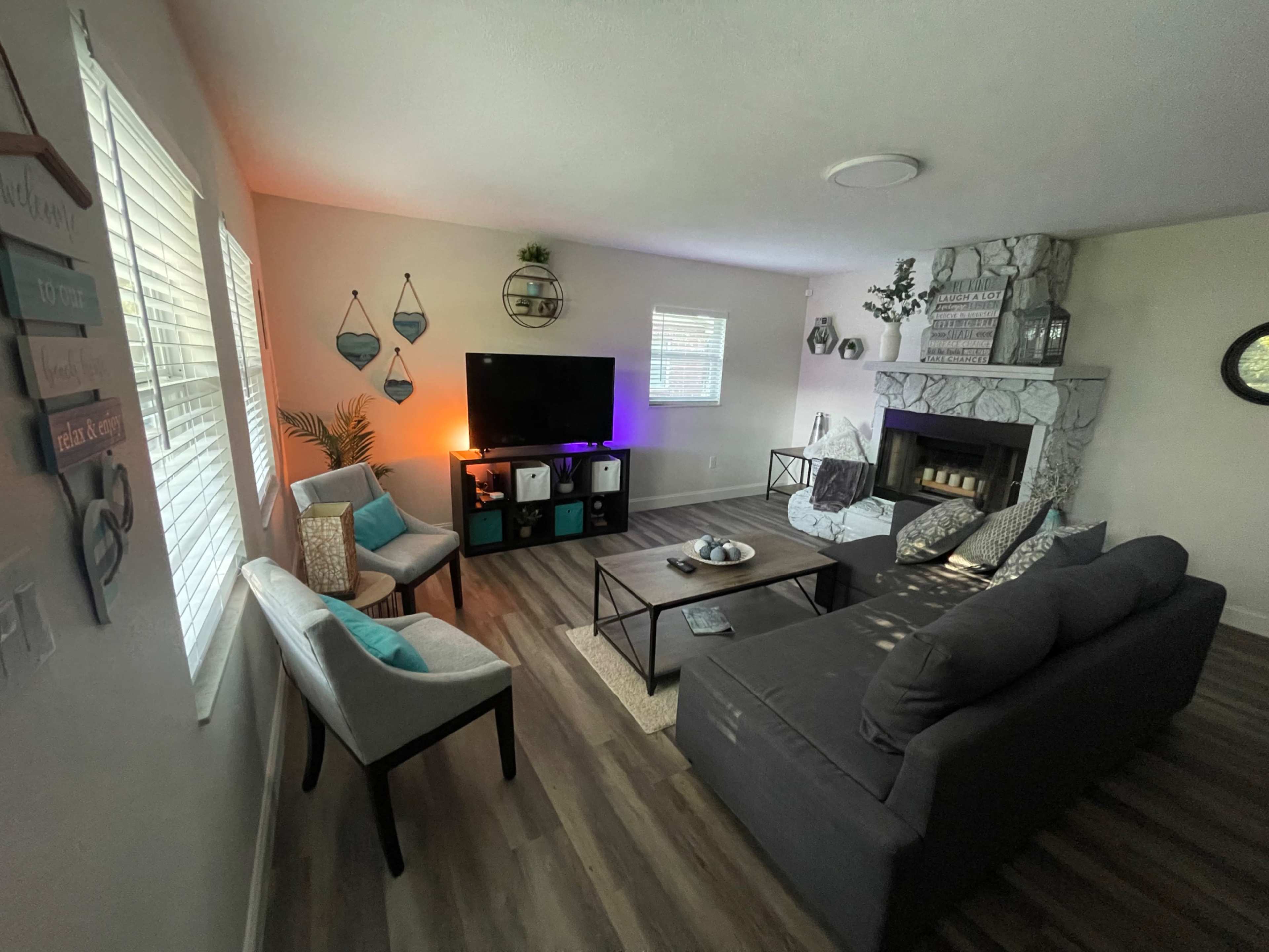 A modern living room with a gray sofa, two light-colored chairs, a television mounted on the wall, and a stone fireplace.