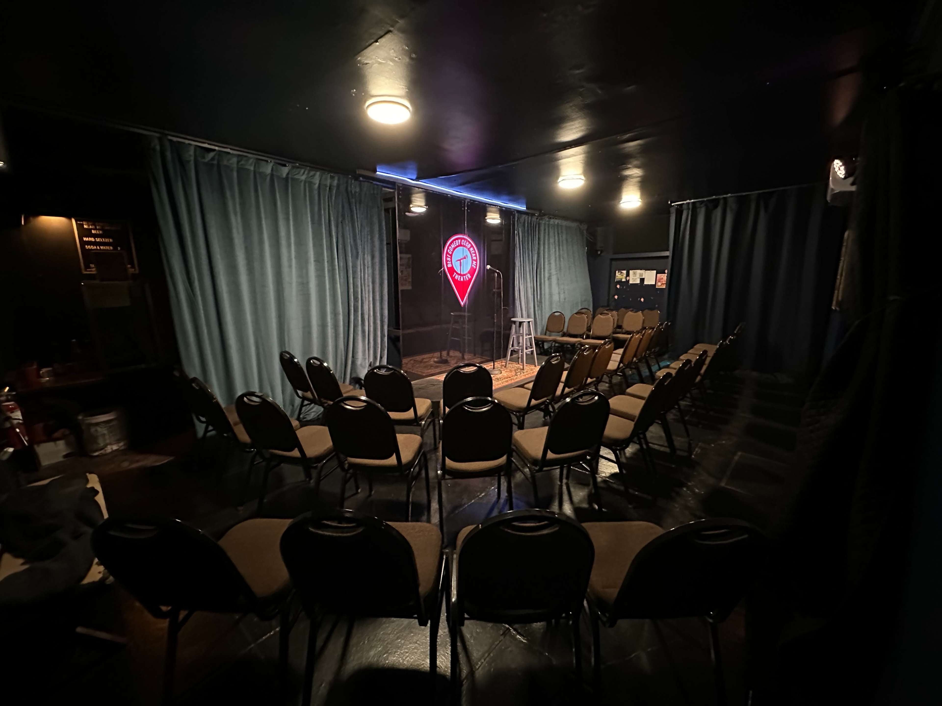 The image shows a small, intimate comedy club setup with circular seating arranged around a stage featuring a microphone and a stool.