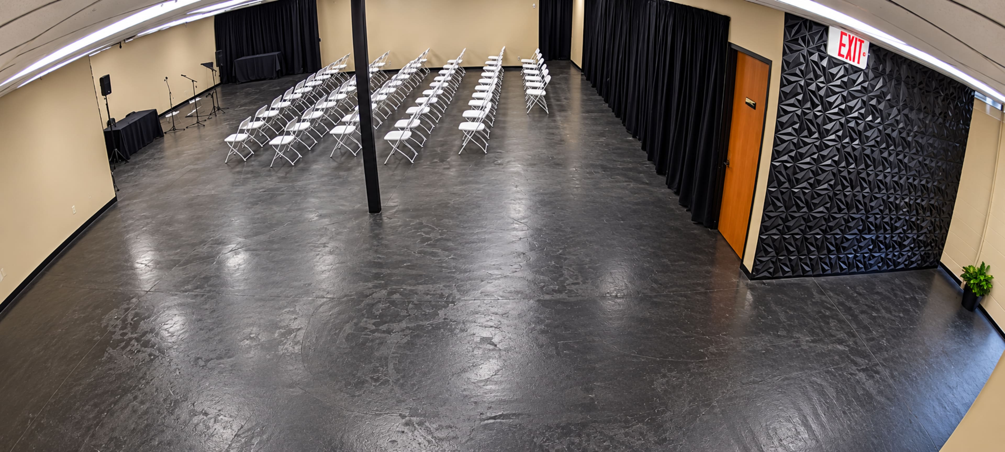 The image depicts a spacious room with rows of white folding chairs arranged facing a black partition and an exit door, featuring a textured wall on one side.