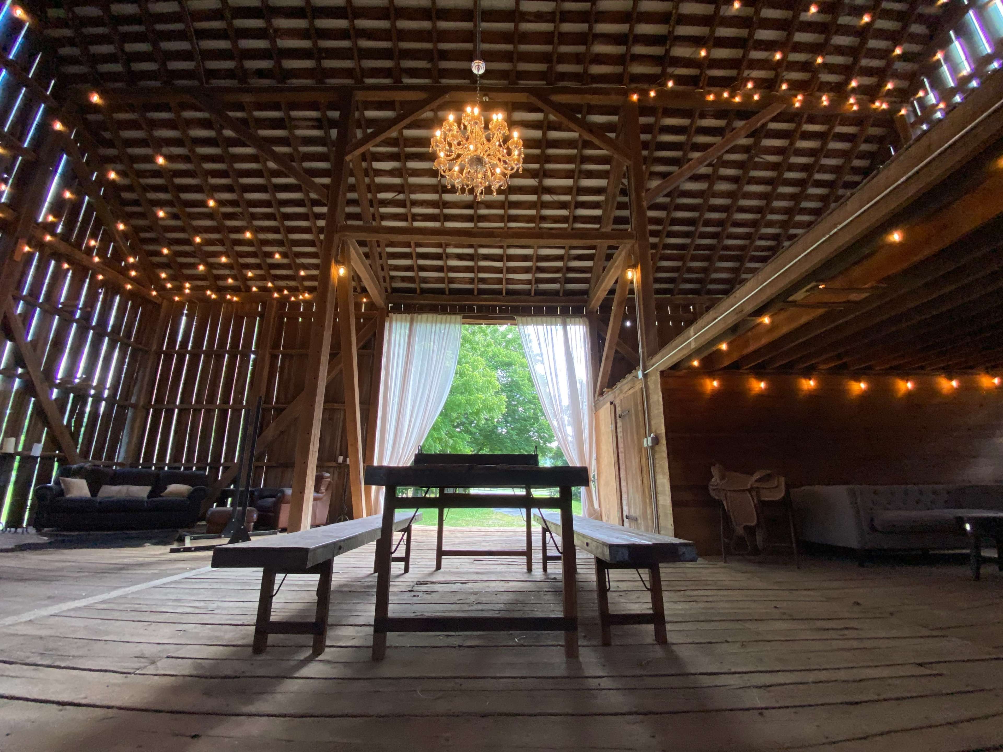 The interior of a barn features wooden beams, a chandelier, and benches arranged around a central space, with soft curtains and string lights enhancing the ambiance.