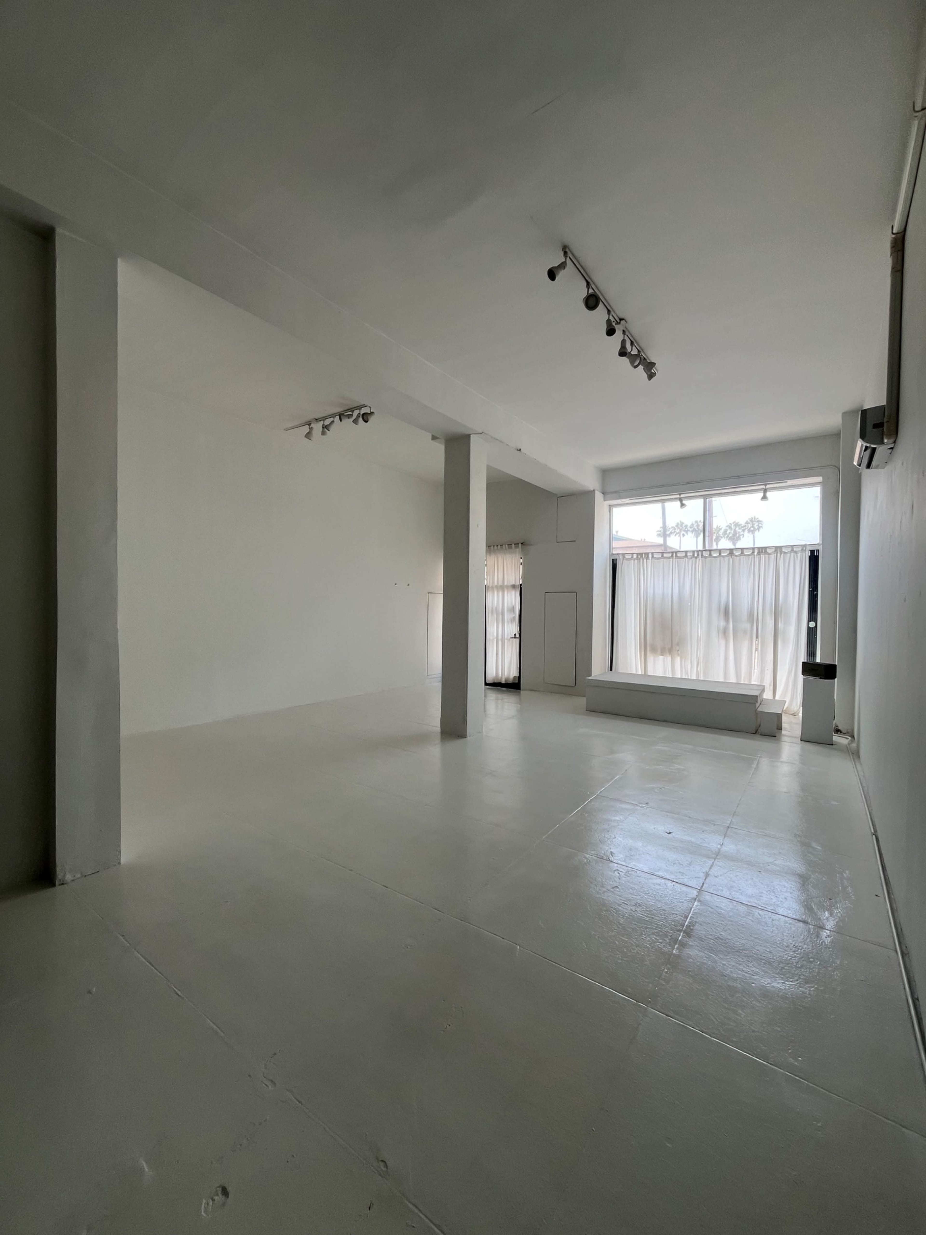 The image shows a spacious, empty room with white walls and a polished floor, featuring large windows dressed in sheer curtains.