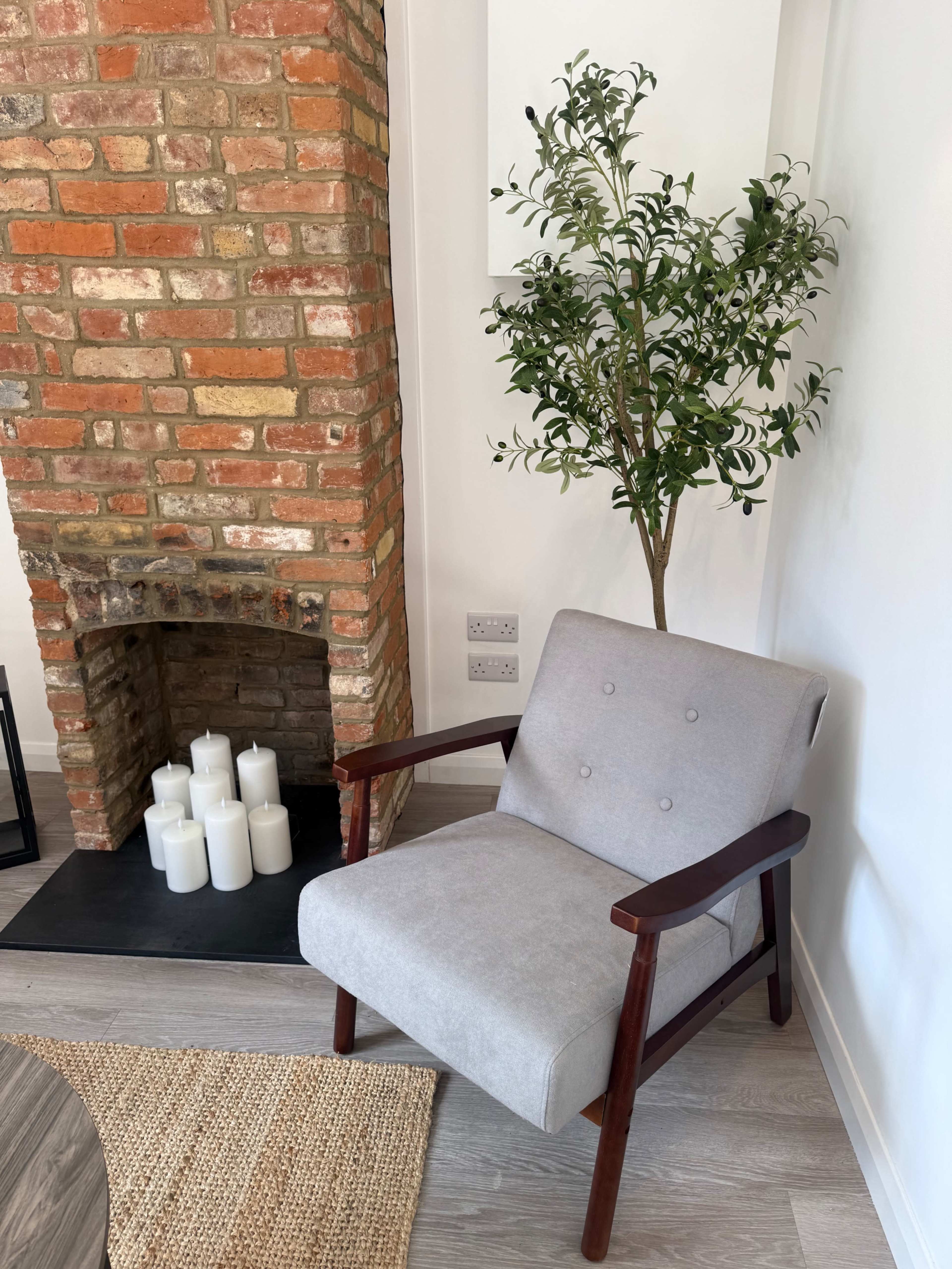 The image displays a gray armchair with wooden arms positioned next to a brick fireplace, which contains a row of white candles and a small green plant nearby.