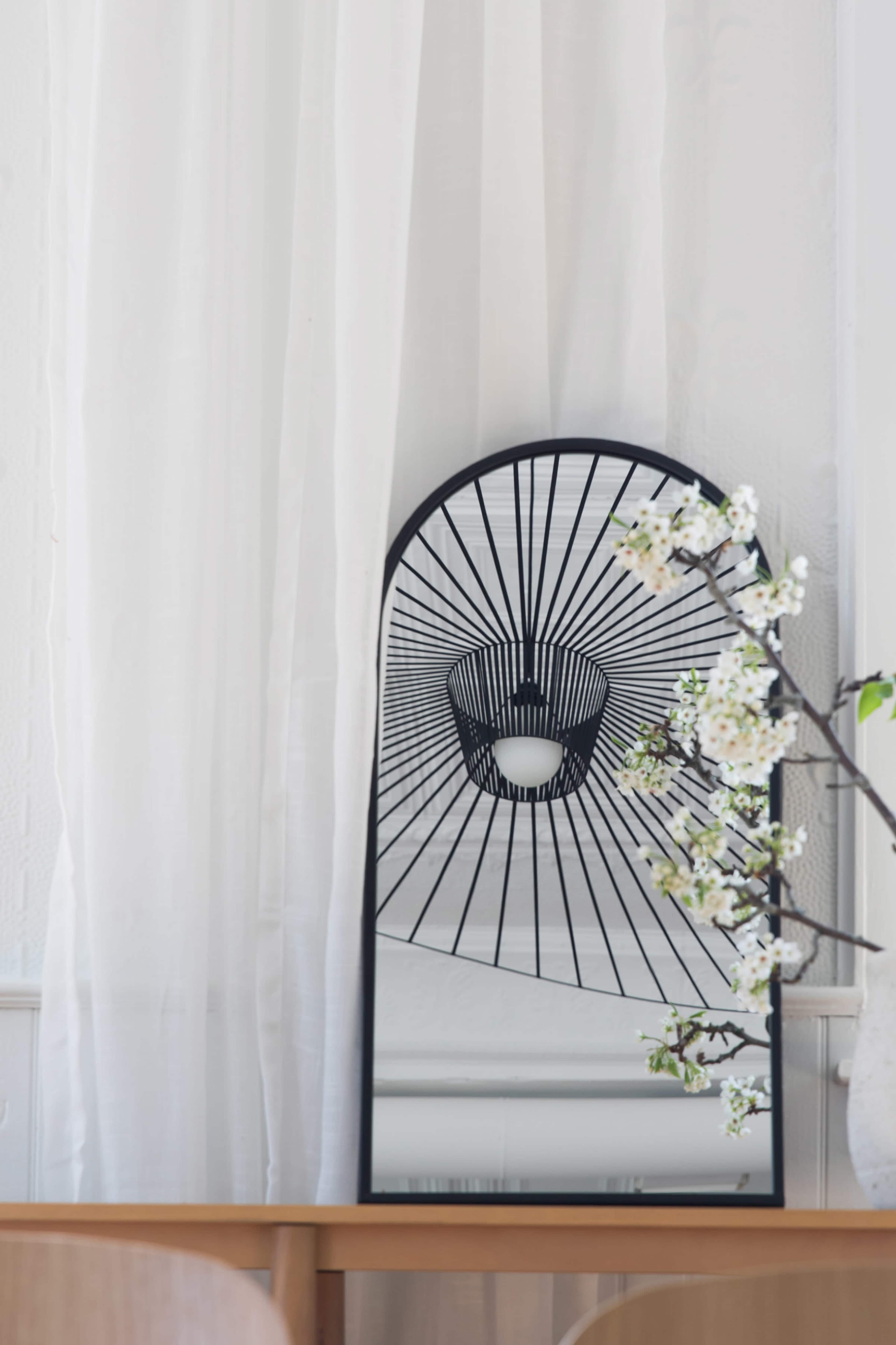 A black decorative mirror with a radial design is positioned against a white wall, reflecting light curtains and a branch with white flowers on a wooden table.