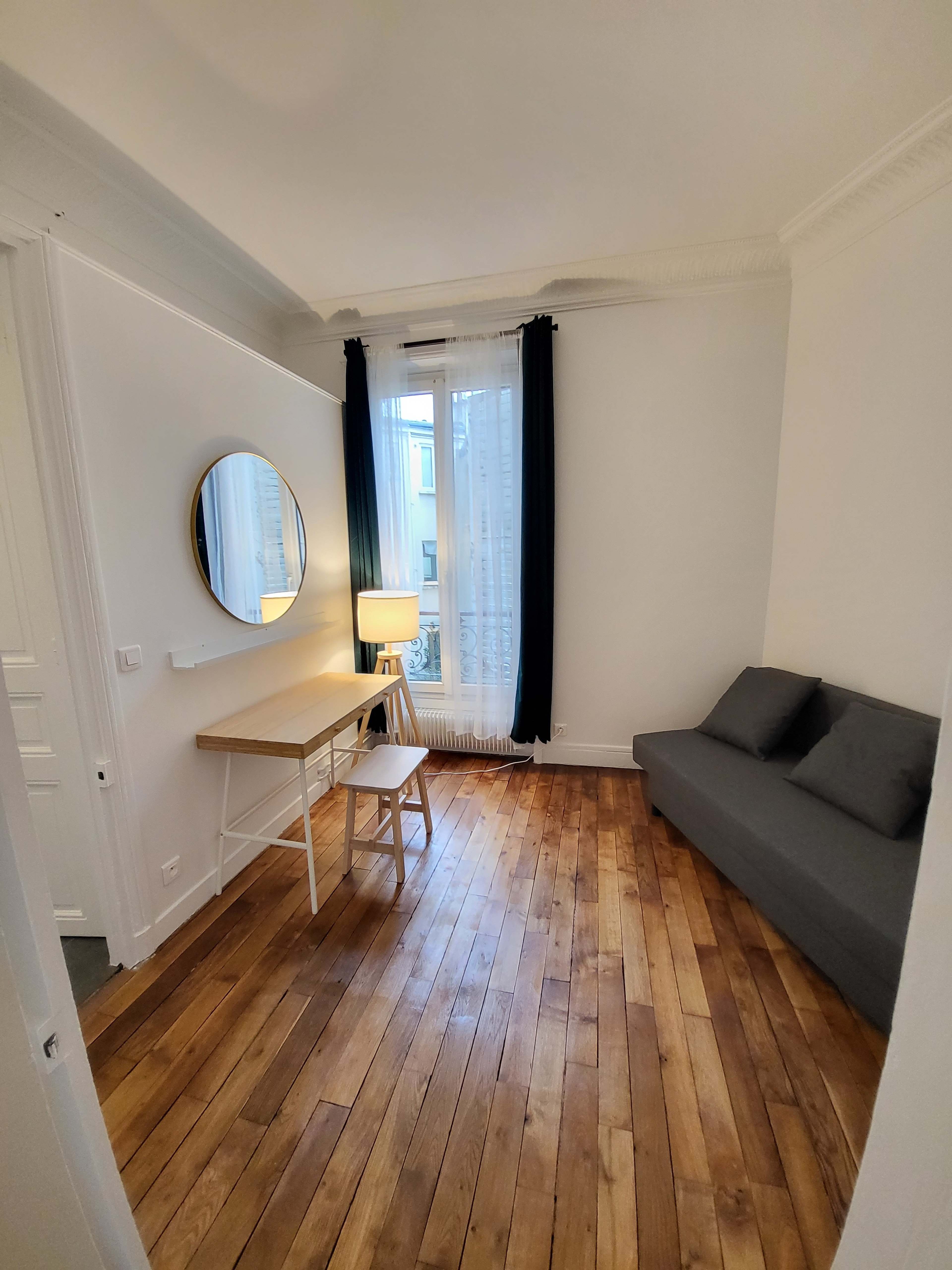 The image shows a small, well-lit room featuring a desk with a chair, a round mirror on the wall, a sofa, and a lamp, all set on wooden flooring.