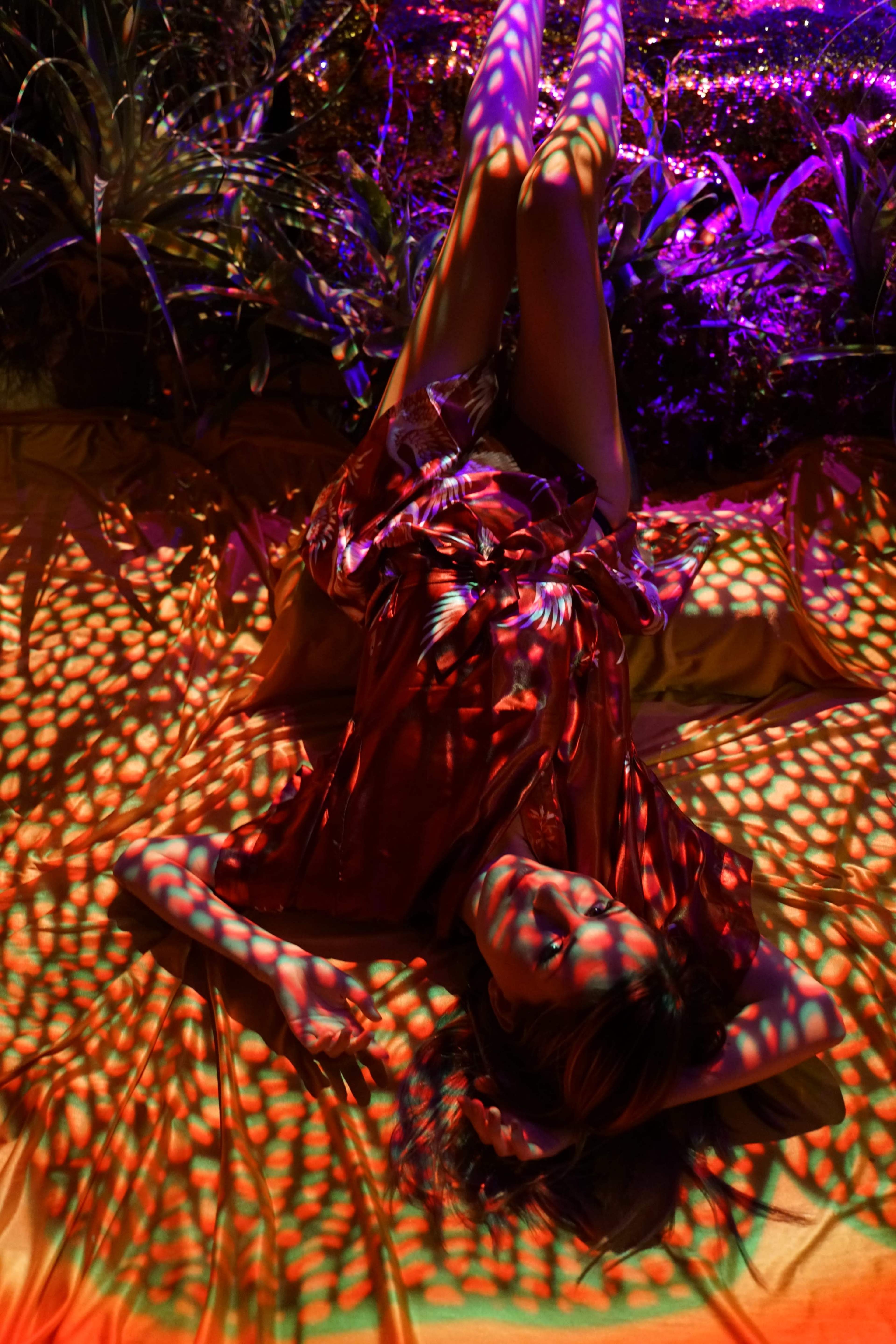 A person lies on a colorful patterned surface surrounded by plants, illuminated by vibrant lights.
