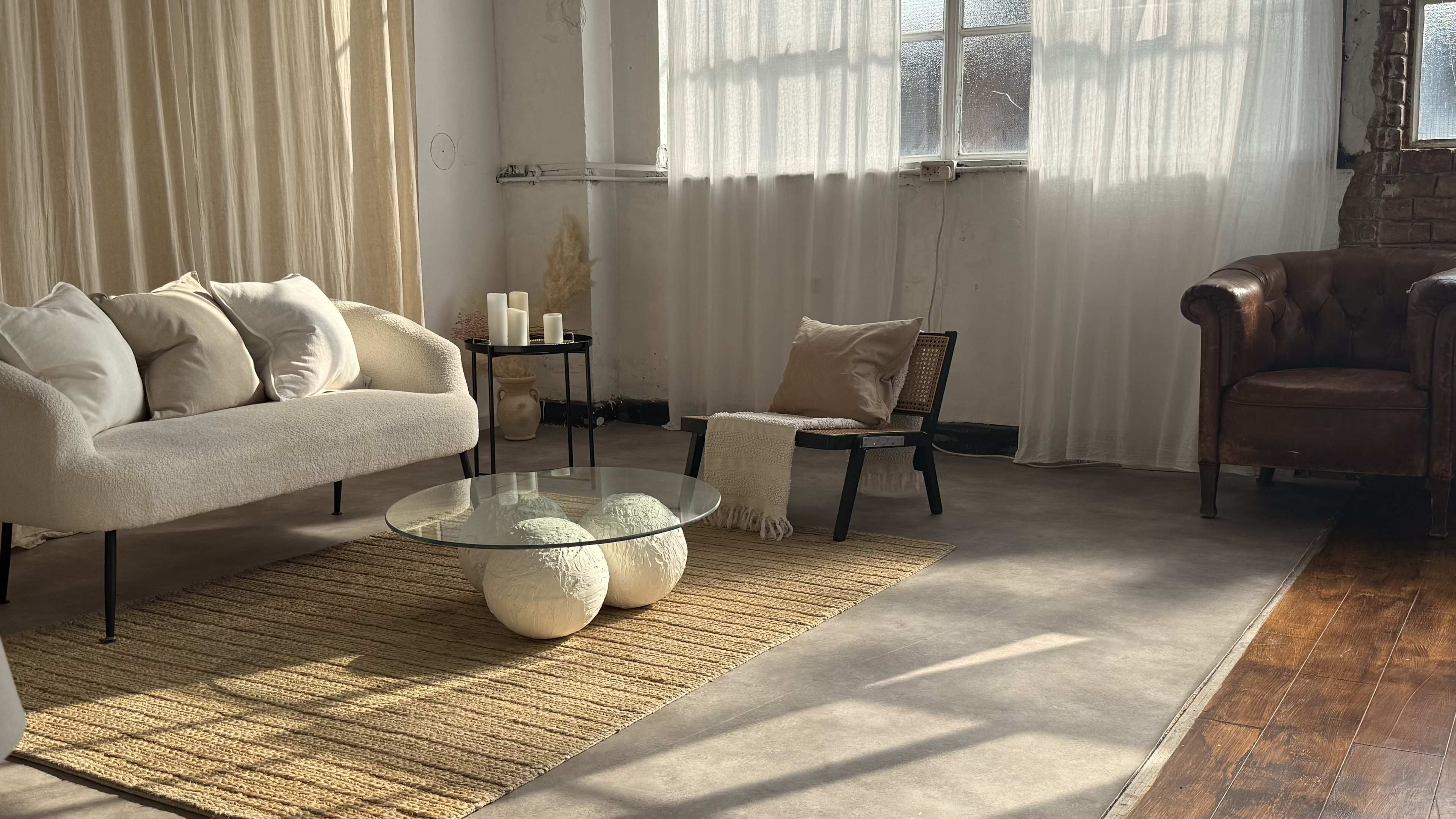 The scene depicts a minimalist living space featuring a sofa, an armchair, a glass coffee table, and natural light flooding through sheer curtains.