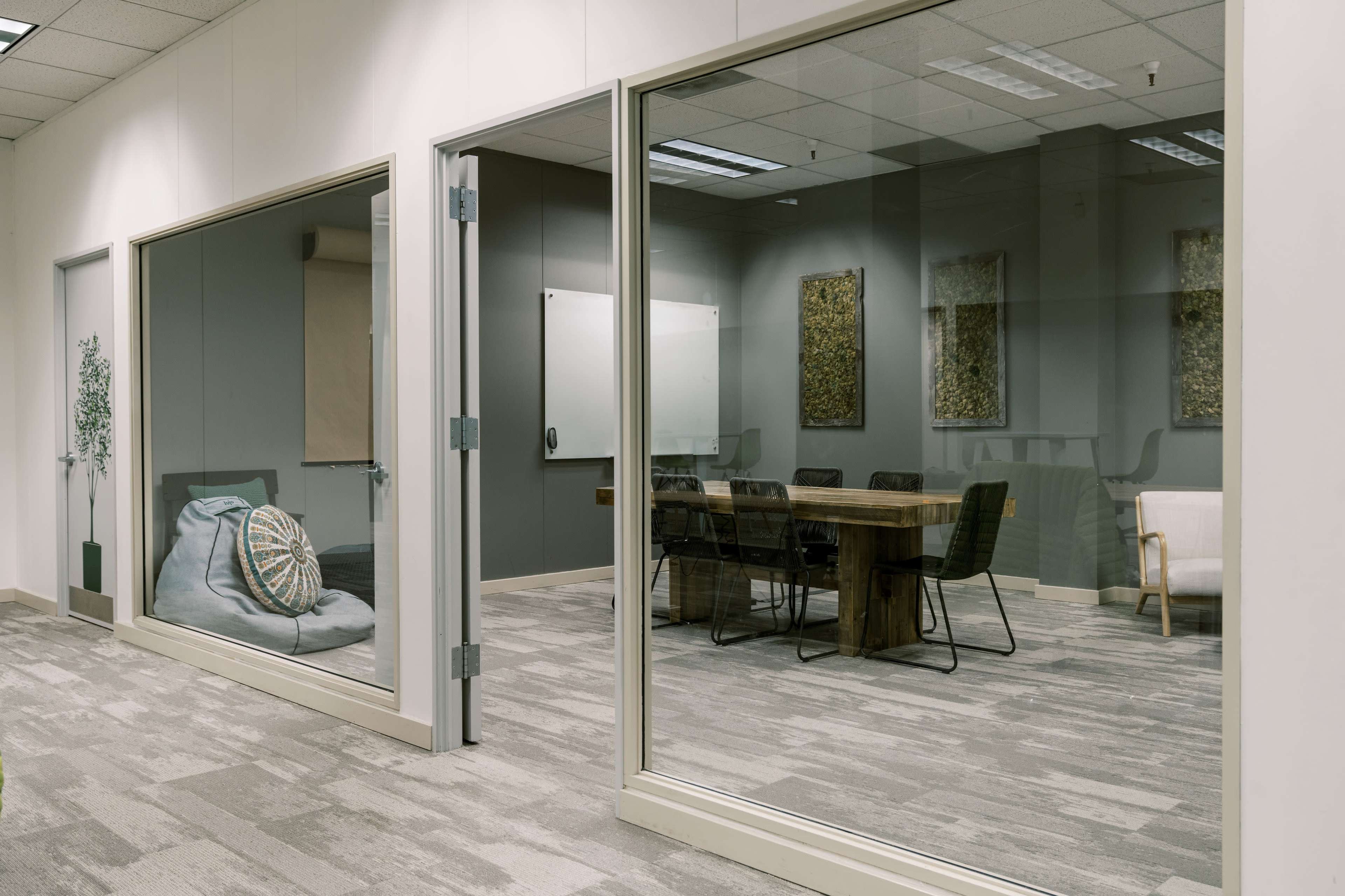 The image shows an office space with two glass-walled rooms, one containing a large wooden table with chairs and the other featuring a bean bag and a plant.