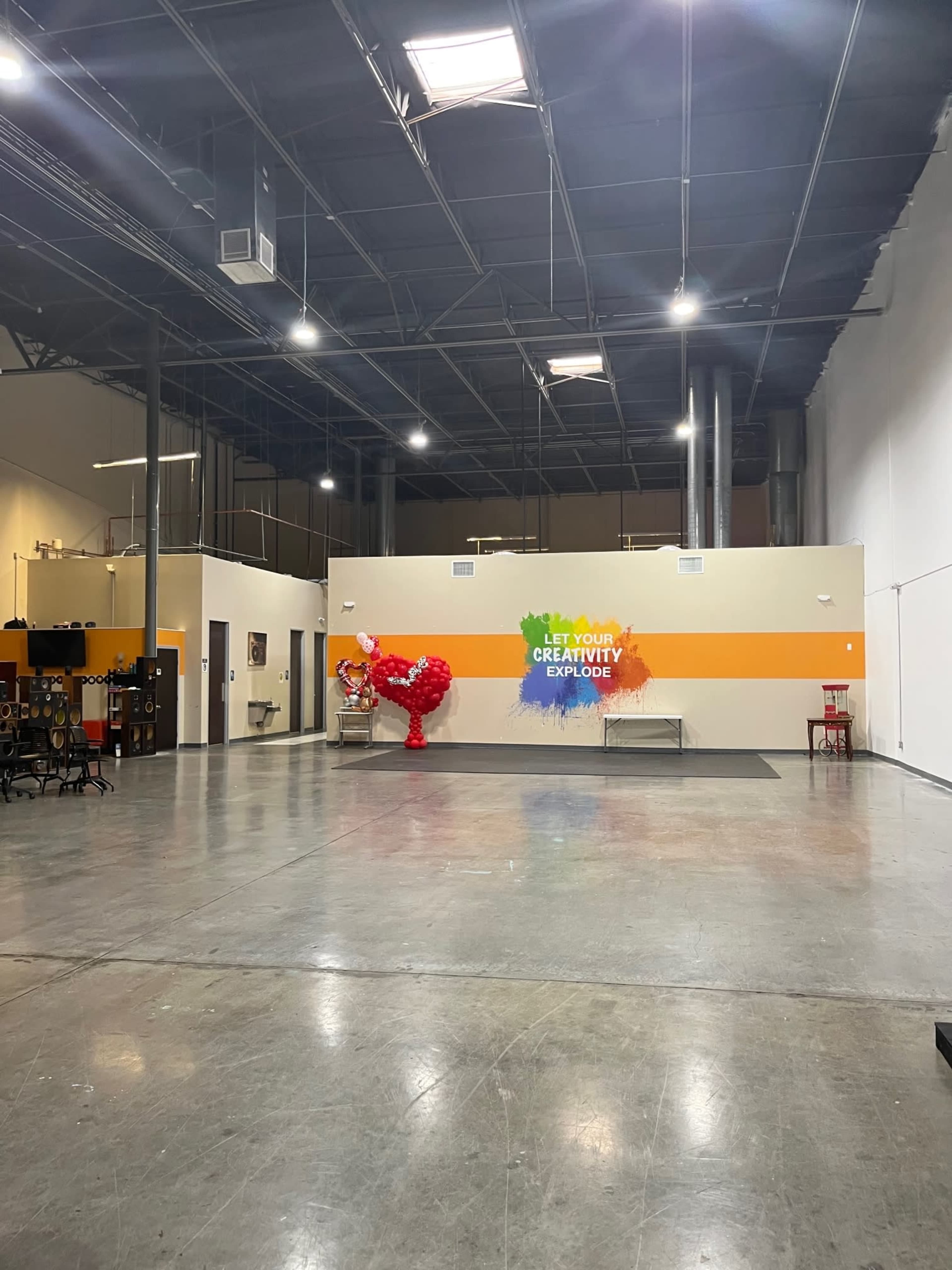 A spacious, empty indoor area with a colorful wall mural that reads "LET YOUR CREATIVITY EXPLODE" and a large balloon sculpture of a rooster in front of it.