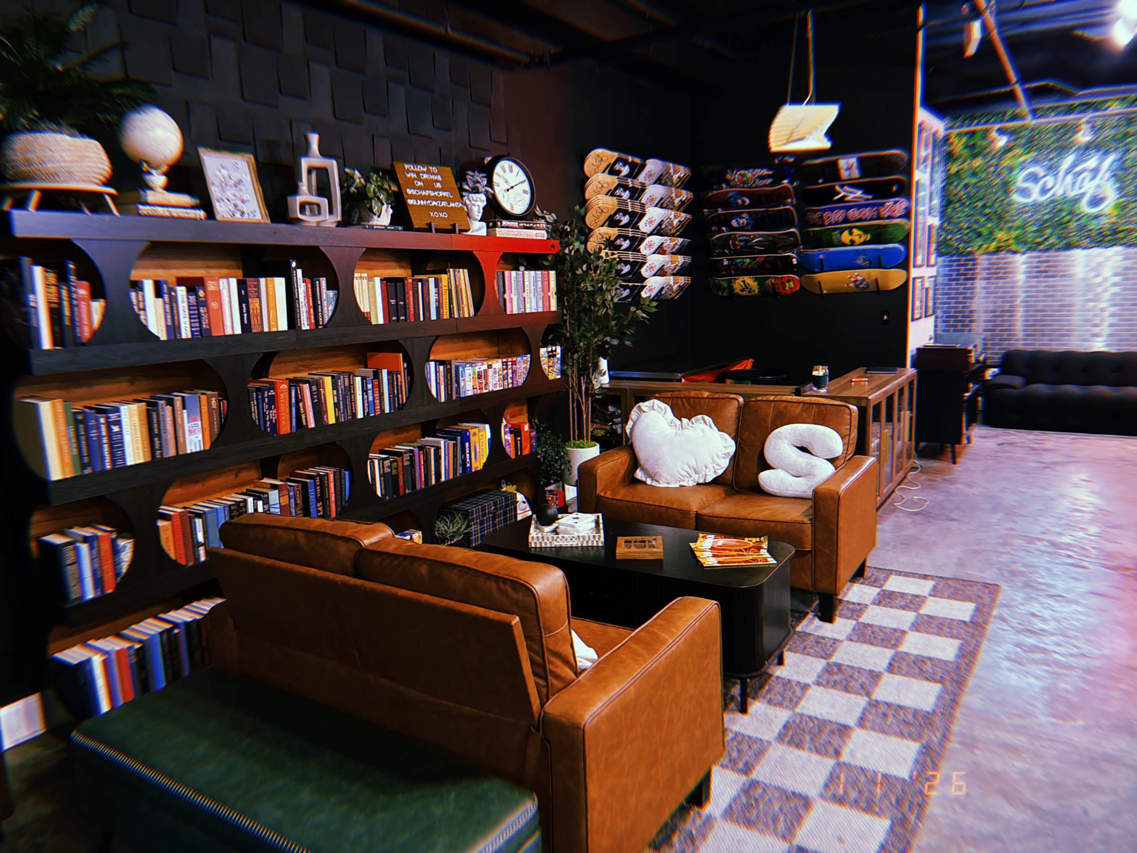 The image shows a cozy lounge area with brown leather sofas, shelves filled with books, a decorative wall clock, and skateboard displays.
